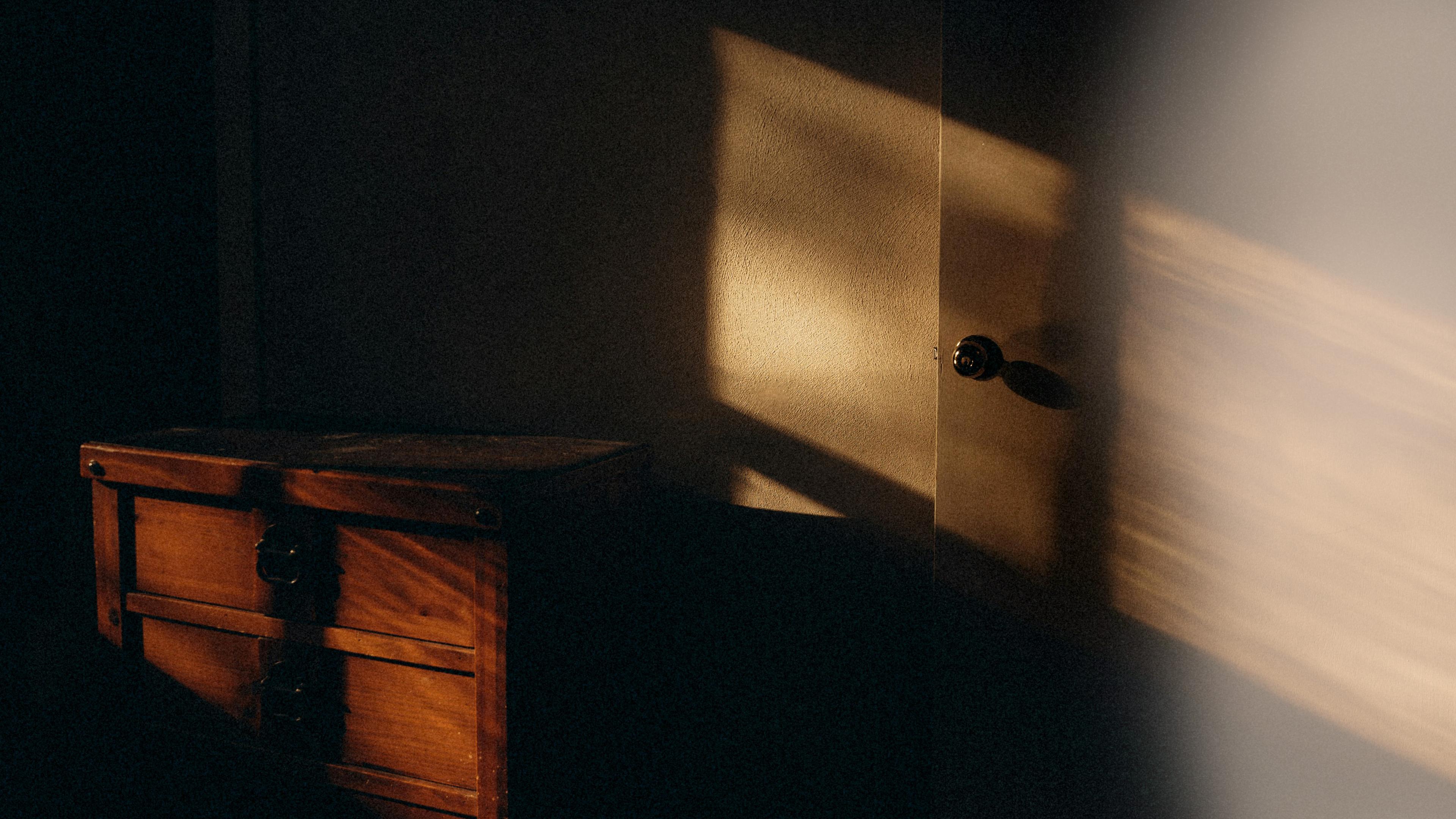 Warm sunlight casting shadows on a wooden chest and a door inside a dimly lit room.
