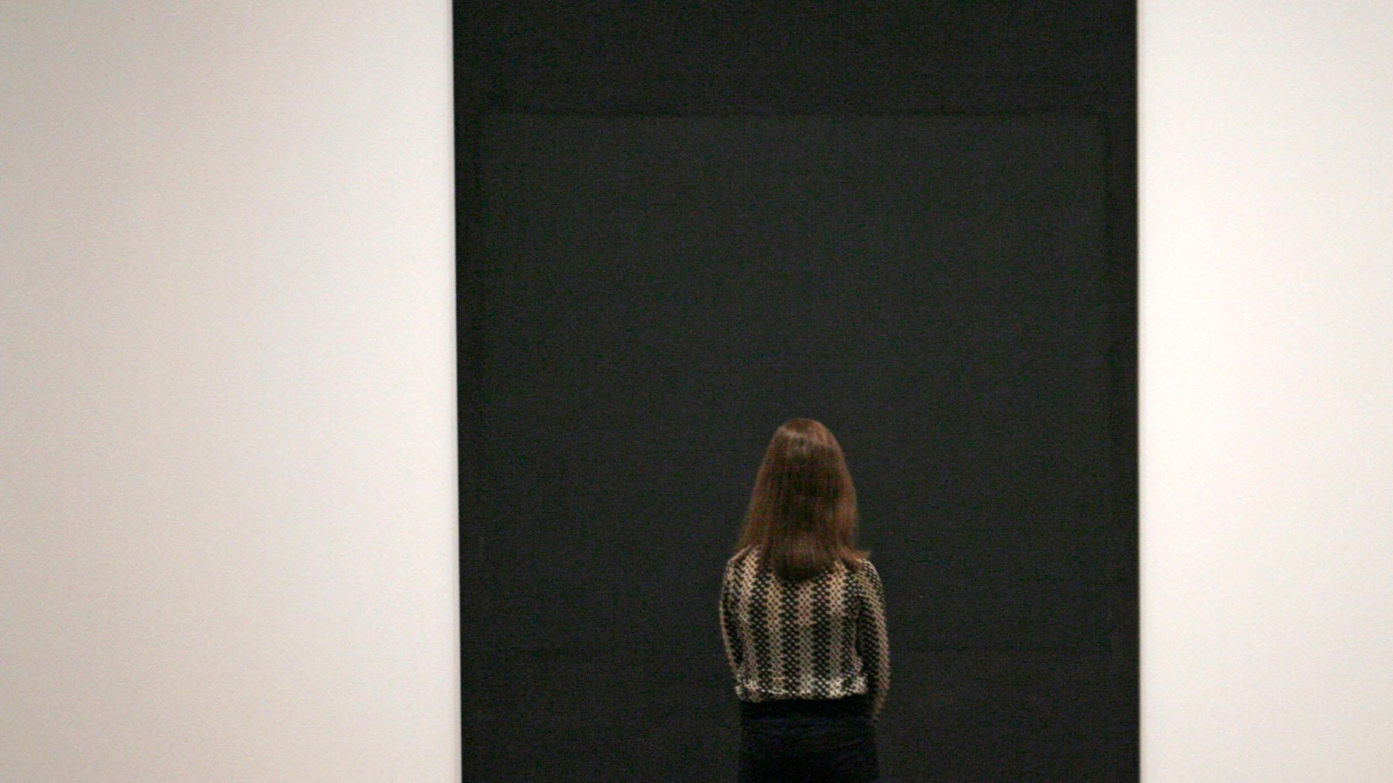 Photo of a woman viewed from behind looking at a large black abstract painting in a minimalist gallery setting.