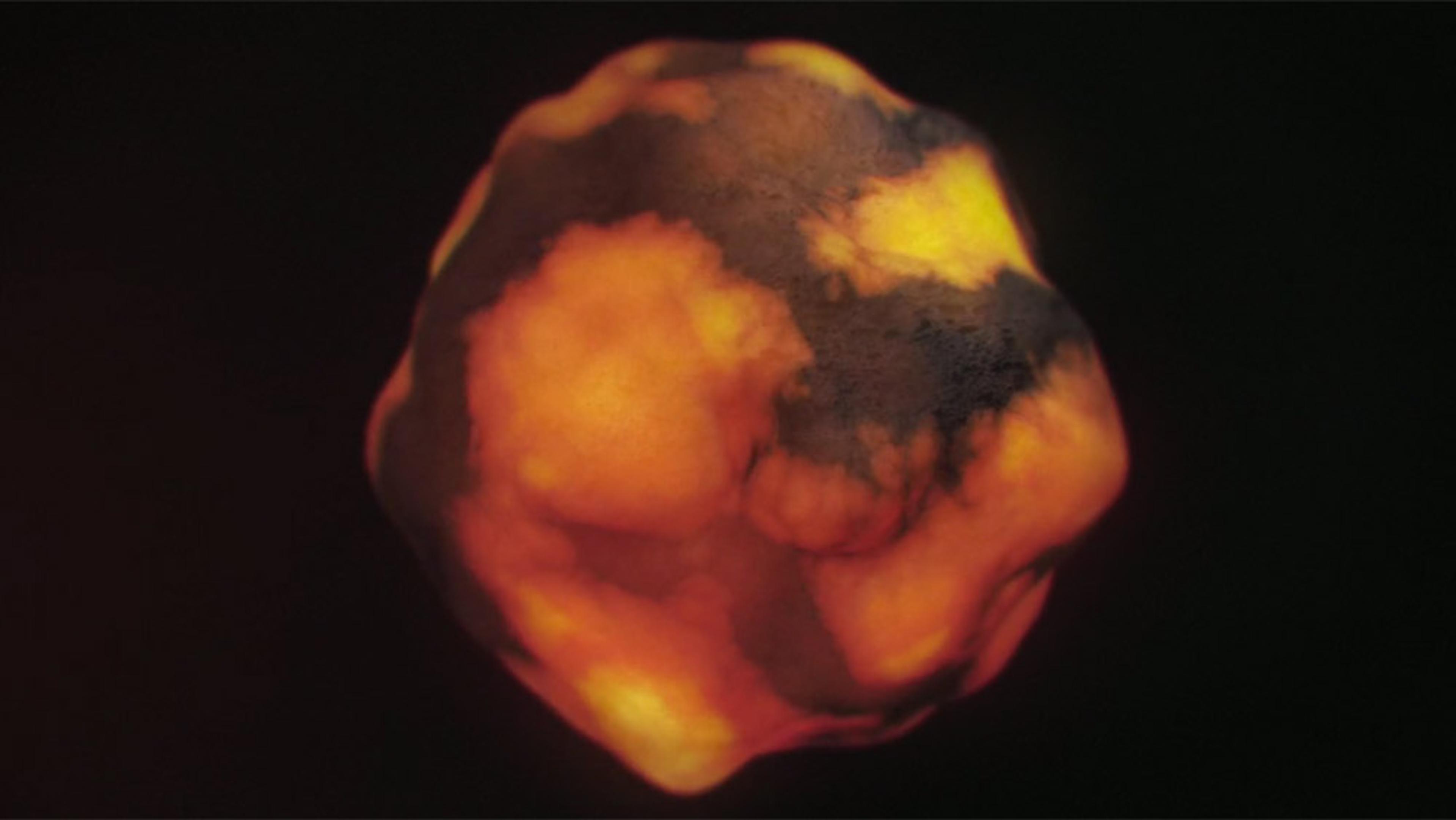 A molten asteroid or space object glowing bright orange with dark patches, set against a black background.