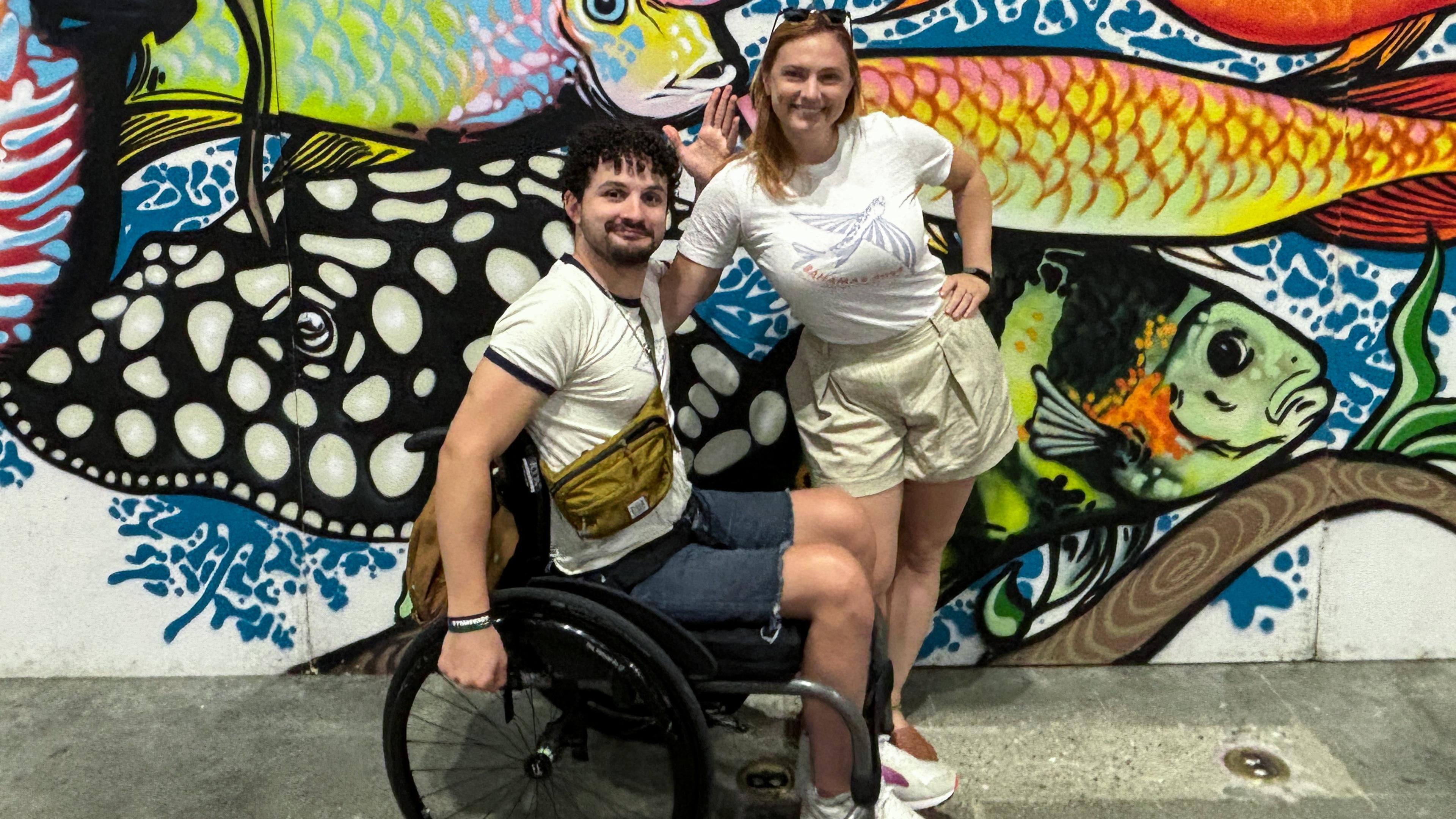 Two people smiling in front of colourful fish graffiti, the man is sitting in a wheelchair the woman is standing beside him.