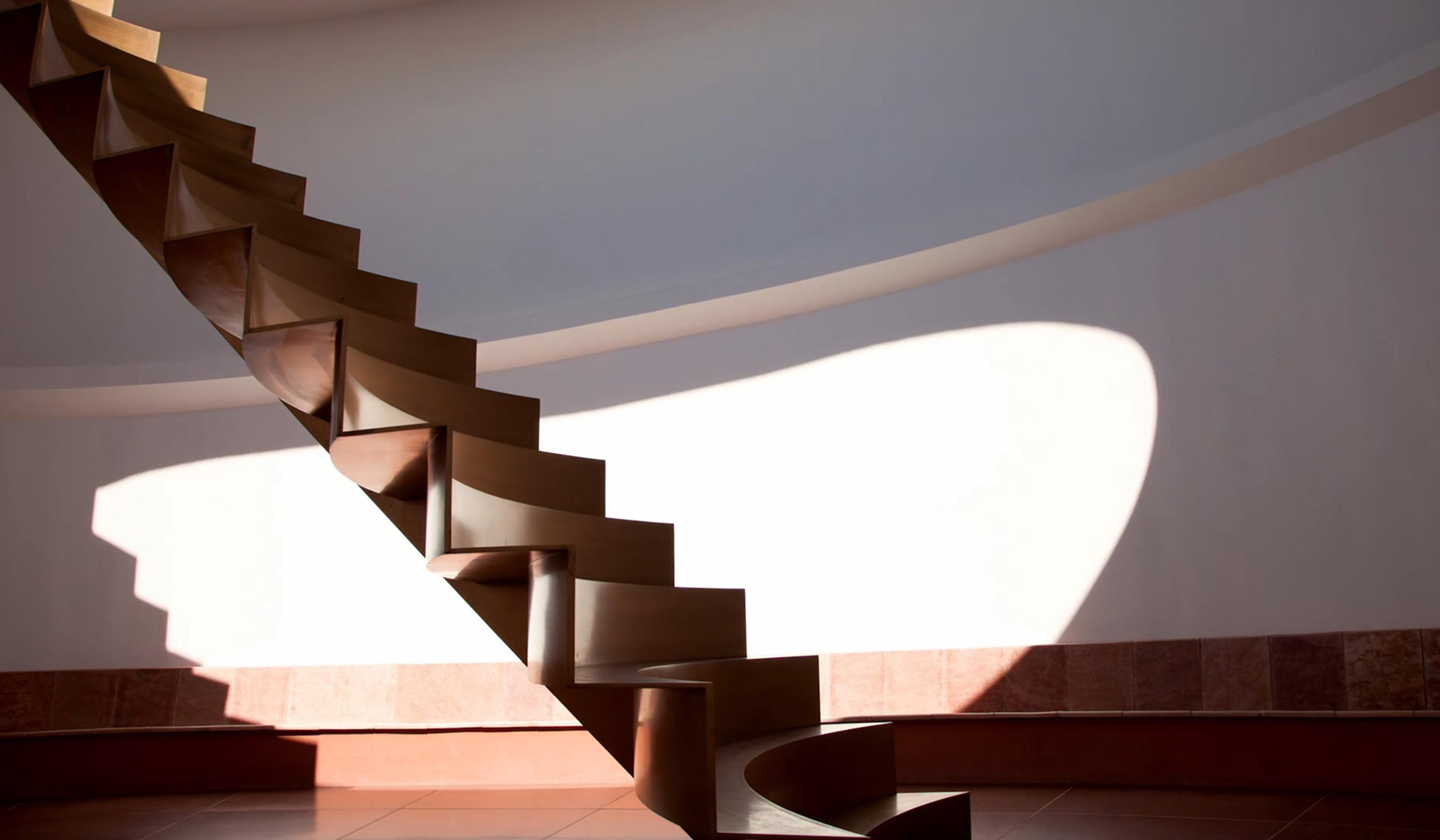 A modern floating staircase casts shadows on a white wall in a minimalist interior with natural light.