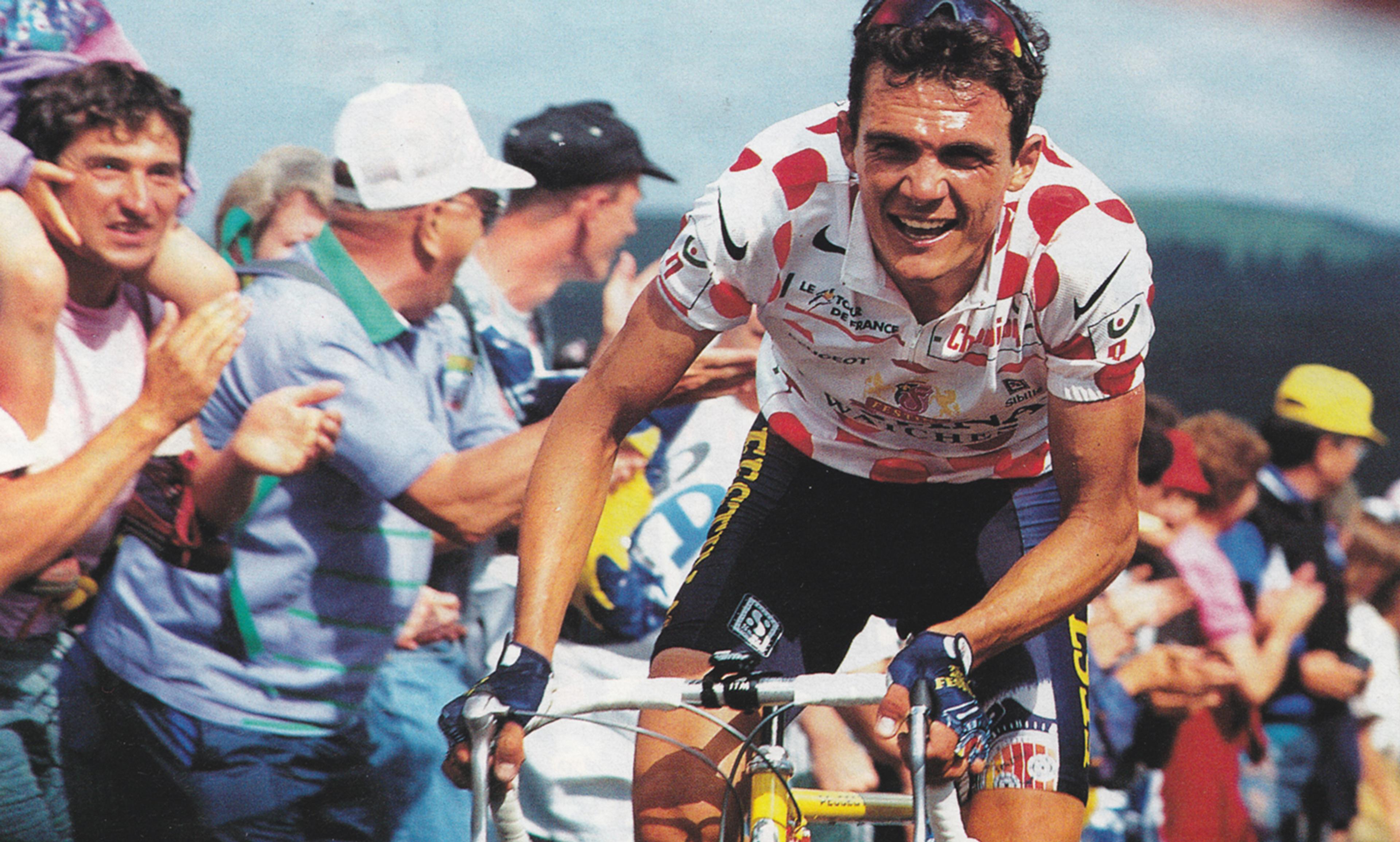 A cyclist wearing a polka dot jersey, riding uphill during a race, cheered by a crowd of spectators.