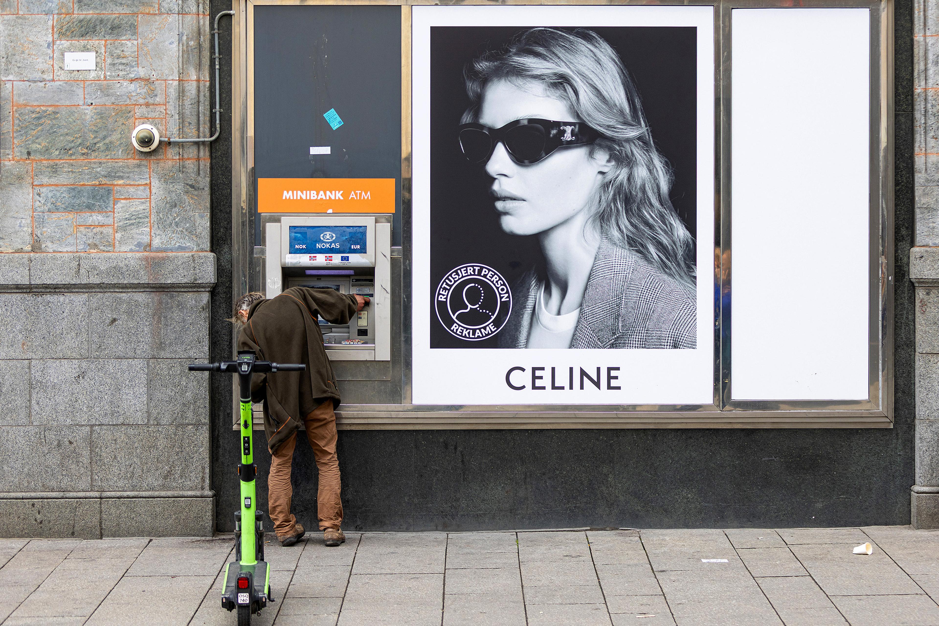 A person using a Minibank ATM next to a Celine advert with an electric scooter parked nearby.