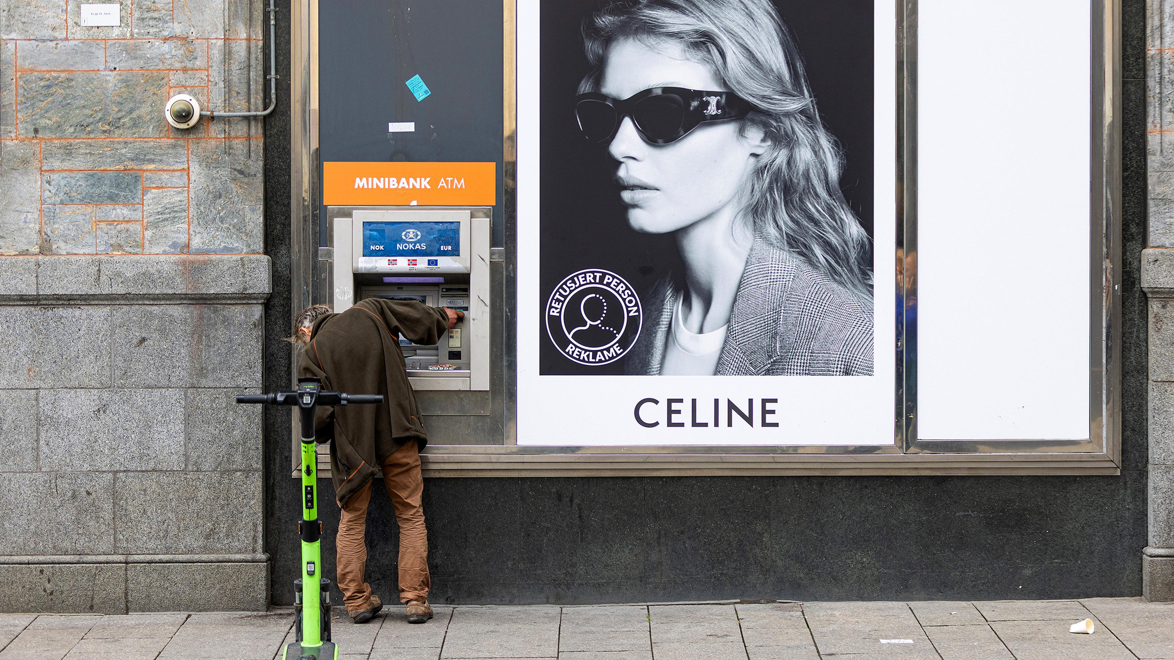 A person using a Minibank ATM next to a Celine advert with an electric scooter parked nearby.