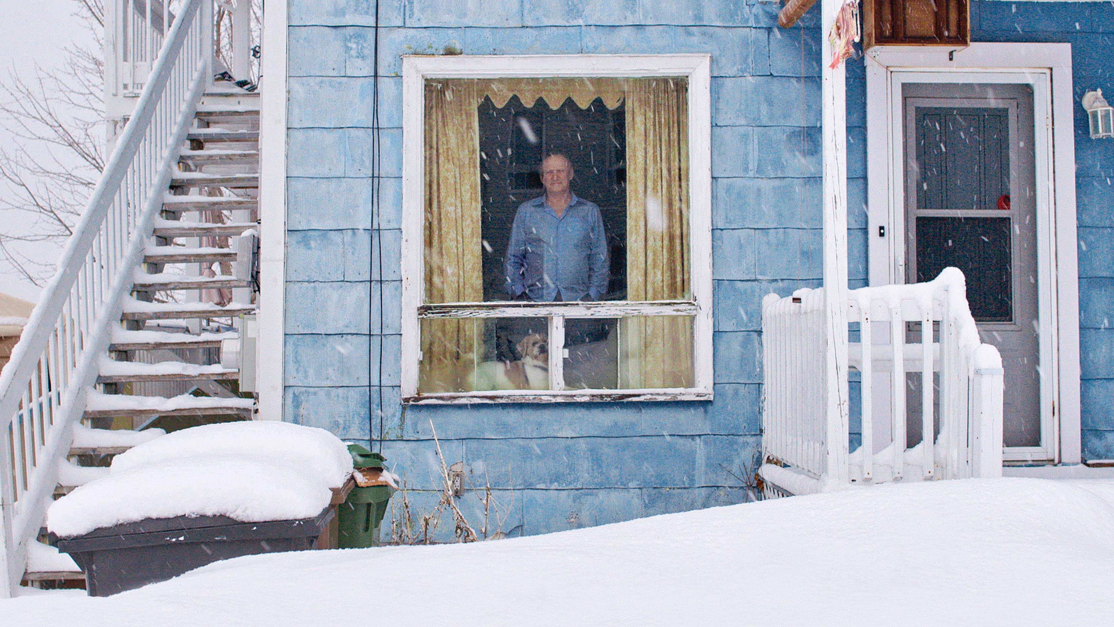 A person and a dog look out from a window of a blue house during snowfall. The house has an external staircase covered with snow on the left, and a snow-covered porch on the right. The scene is set on a snowy day, with visible accumulation on the ground and surfaces.