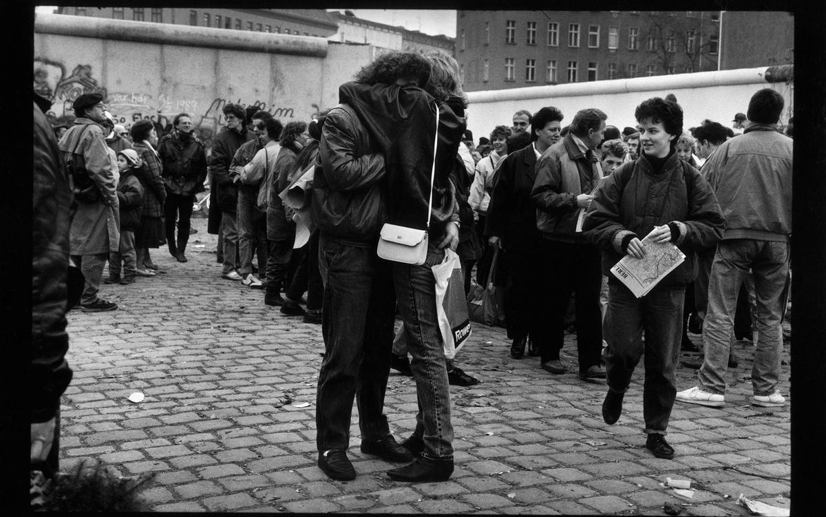 Black and white photo of people gathered by the Berlin Wall, a couple embracing in the foreground.