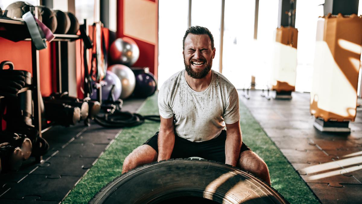 A man lifting a tyre in a gym, showing effort. Fitness equipment and exercise balls are visible in the background.