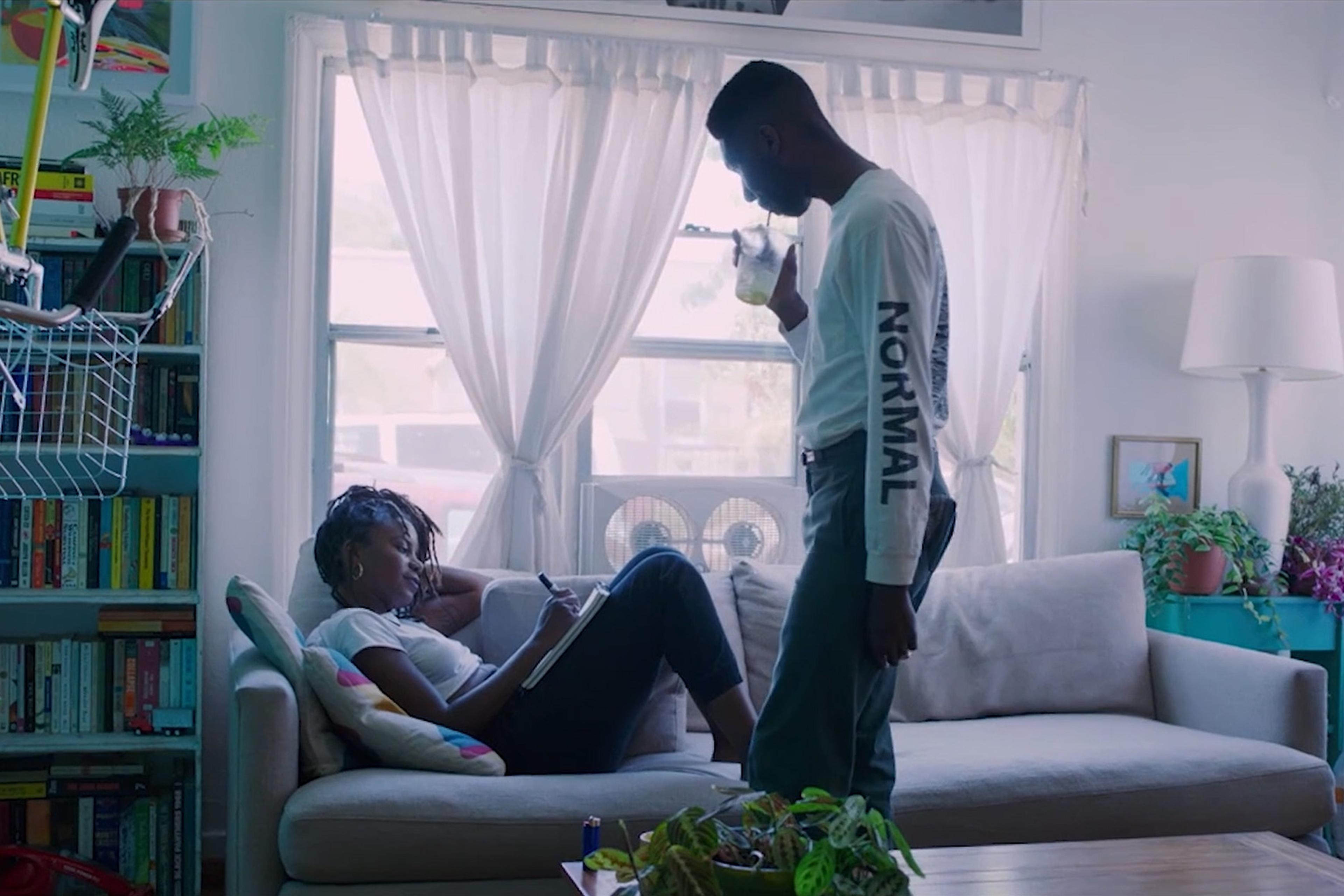 A woman reclines on a sofa writing in a notebook while a man stands nearby drinking a beverage in a brightly lit room with books and plants.