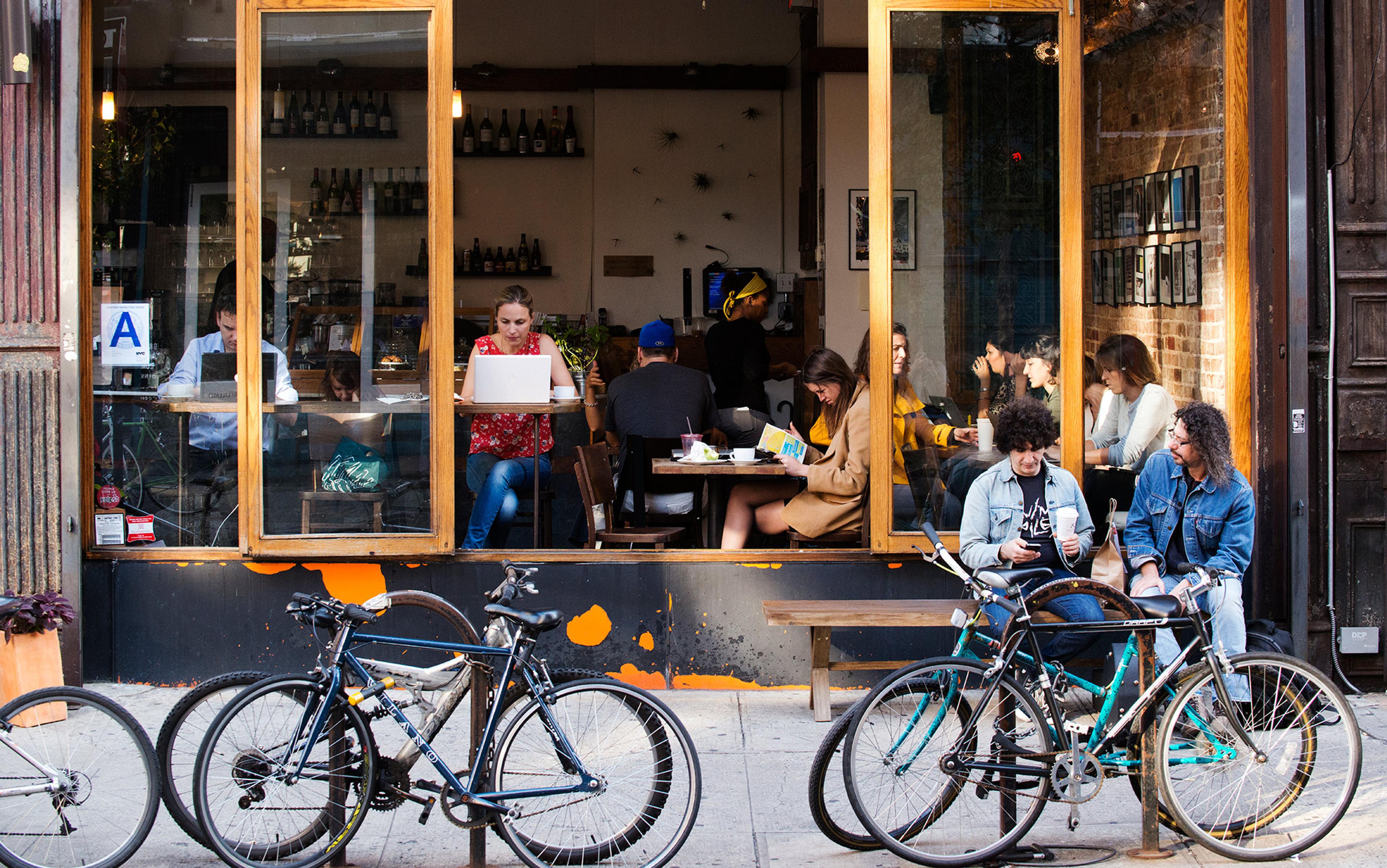 A café with people inside and outside, some working on laptops, some talking, bicycles parked in front on a city street.