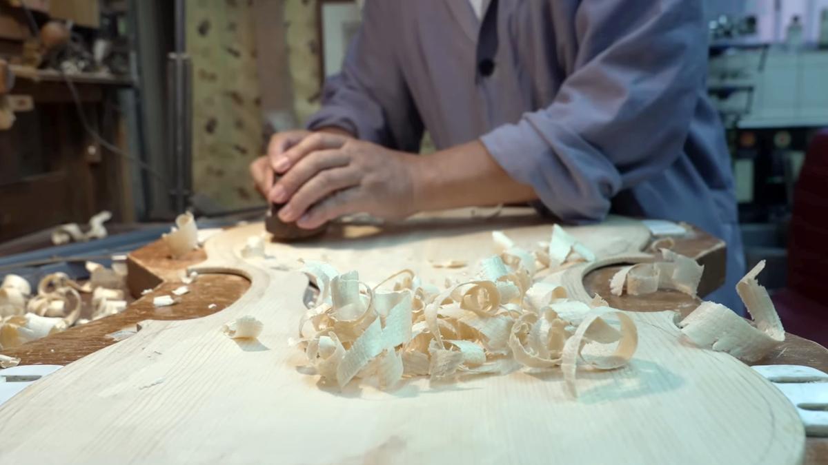 A person in a workshop uses a tool to smooth wood. Wood shavings are scattered on the workbench.