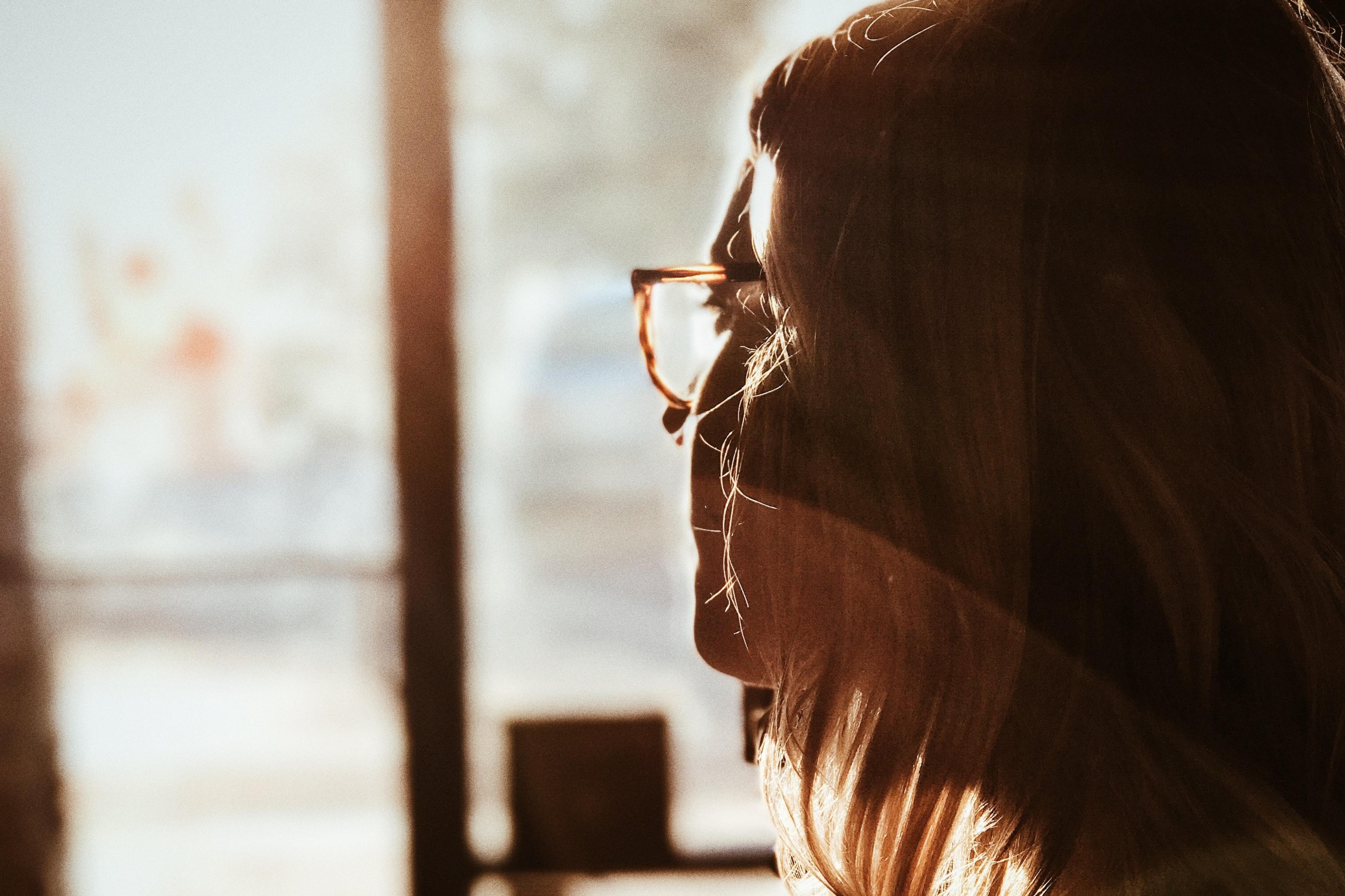 Photo of a woman in profile with glasses, backlit by warm sunlight through a window, creating a soft, serene atmosphere.