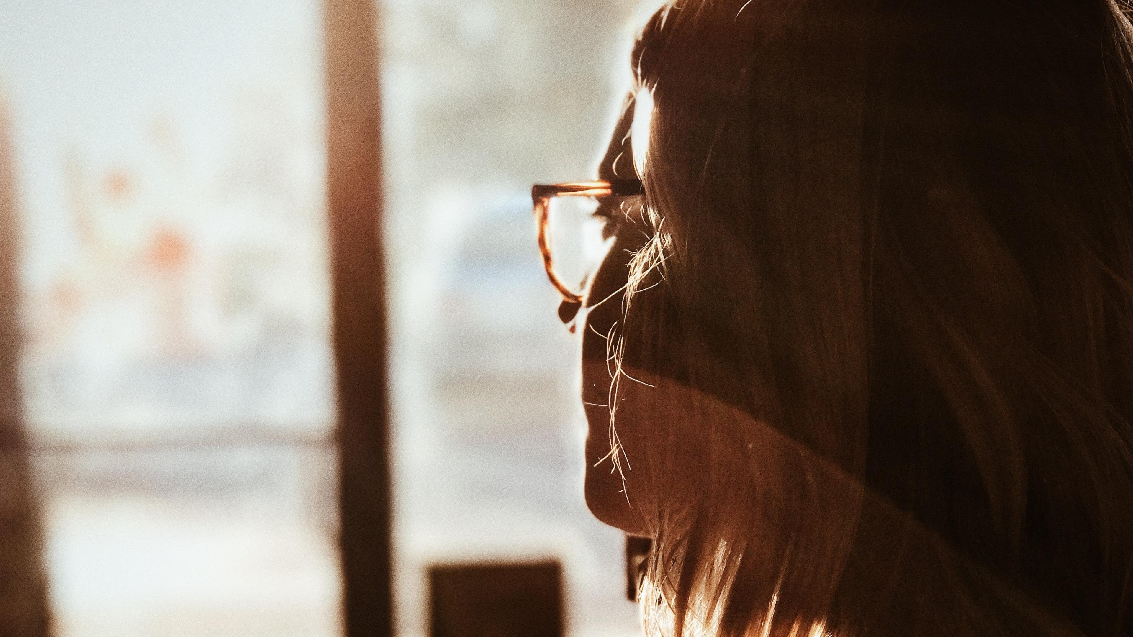 Photo of a woman in profile with glasses, backlit by warm sunlight through a window, creating a soft, serene atmosphere.