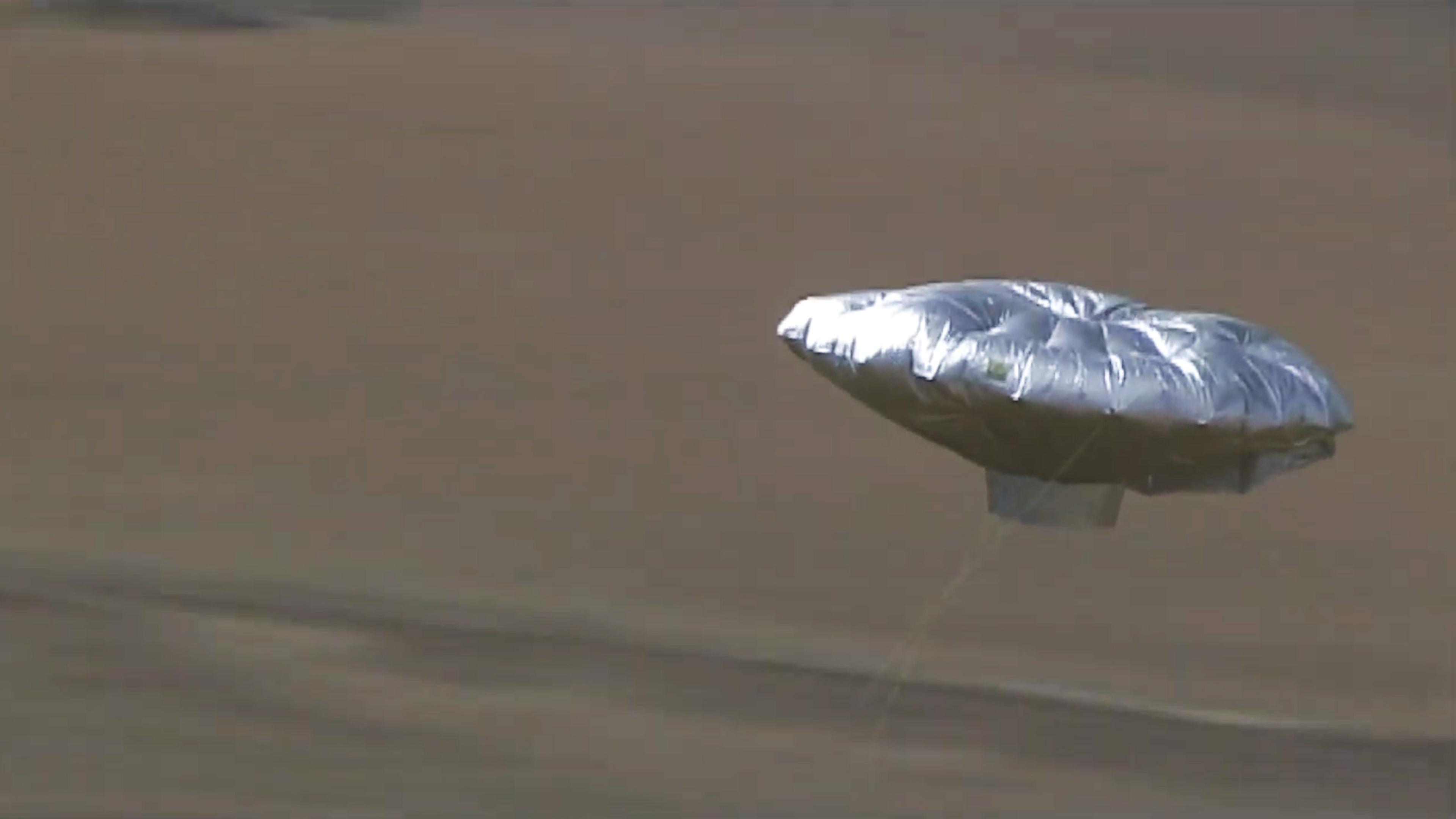 Photo of a silver inflatable balloon floating in the sky over a blurred brownish background, resembling a flying saucer.