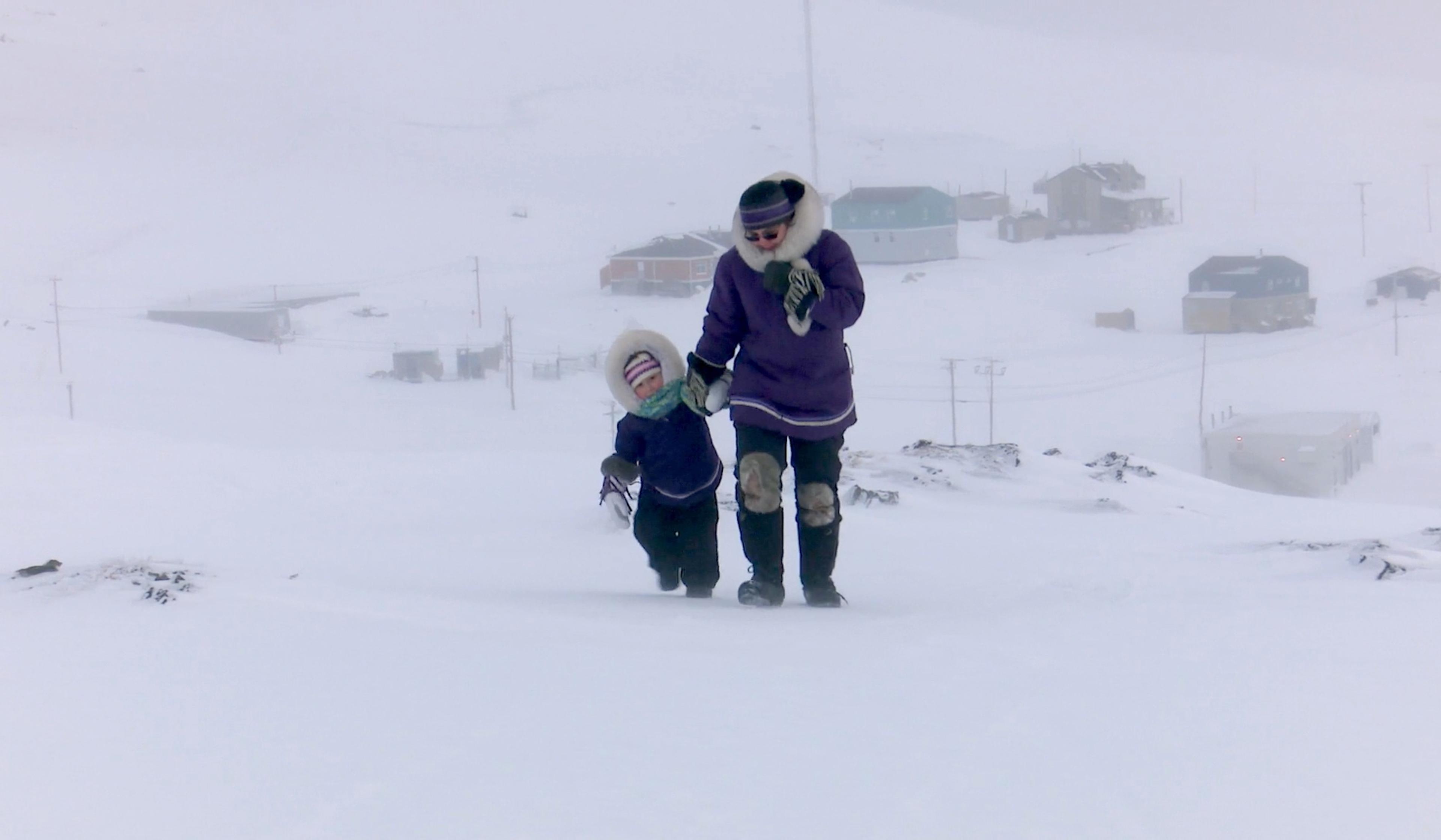 Two people in winter clothing walk through a snowy landscape, with buildings and power lines in the background under heavy snowstorm conditions.