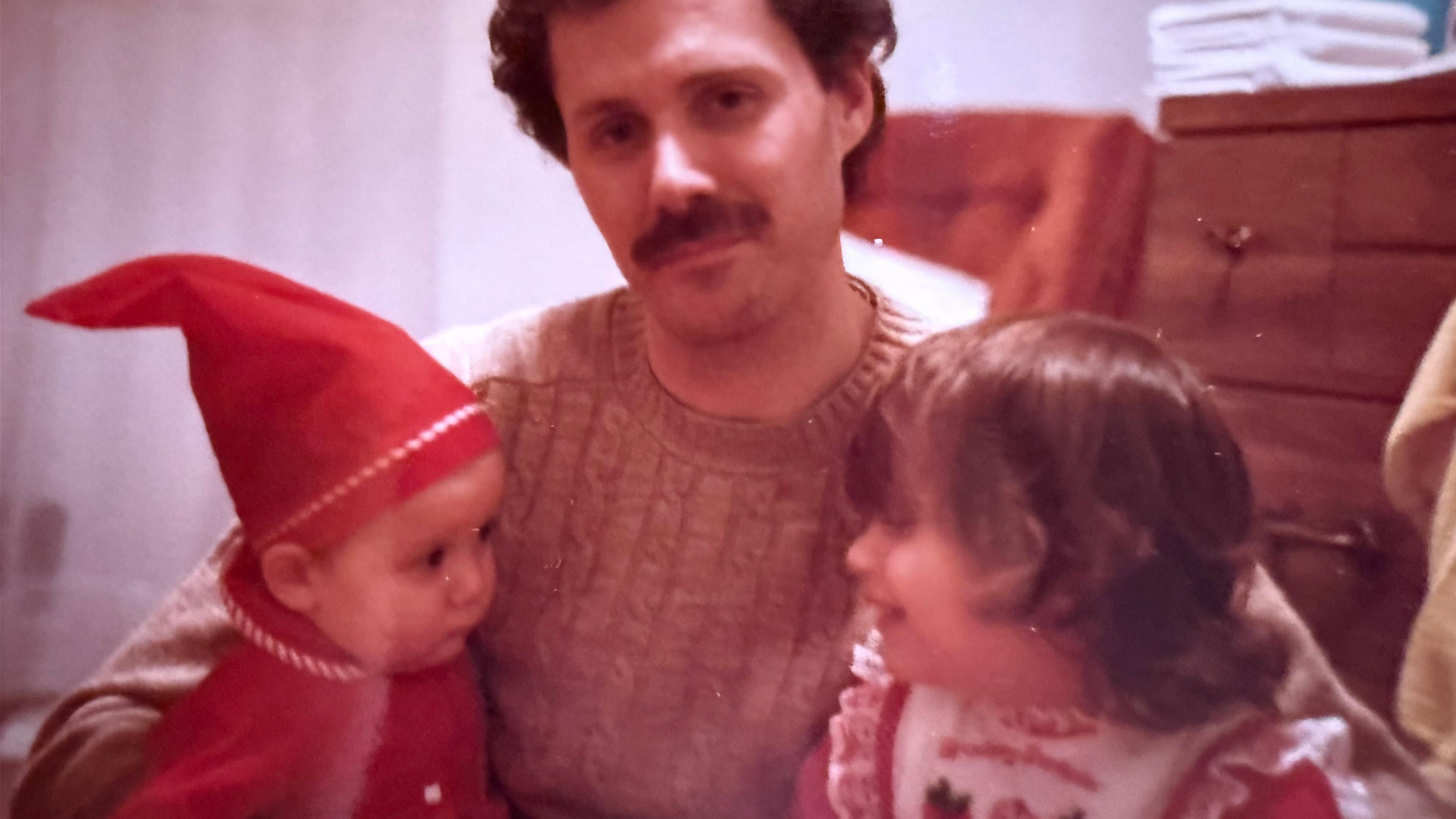 Vintage photo of a man with a moustache holding a baby wearing a red pointed cap and a young child