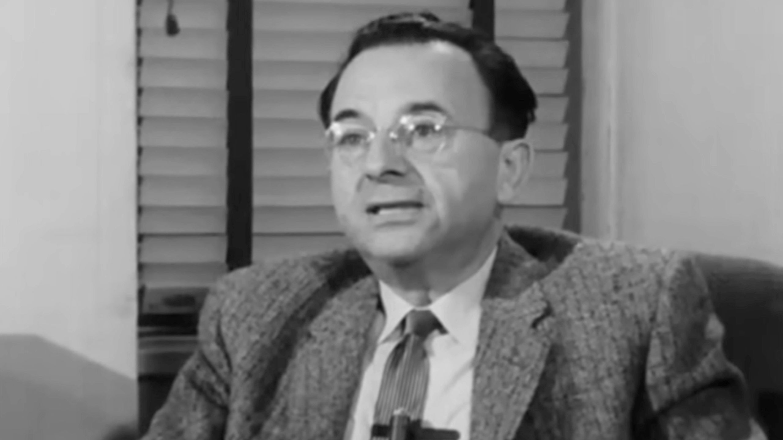 Black and white photo of a man in glasses and a suit speaking, seated indoors with window blinds in the background.