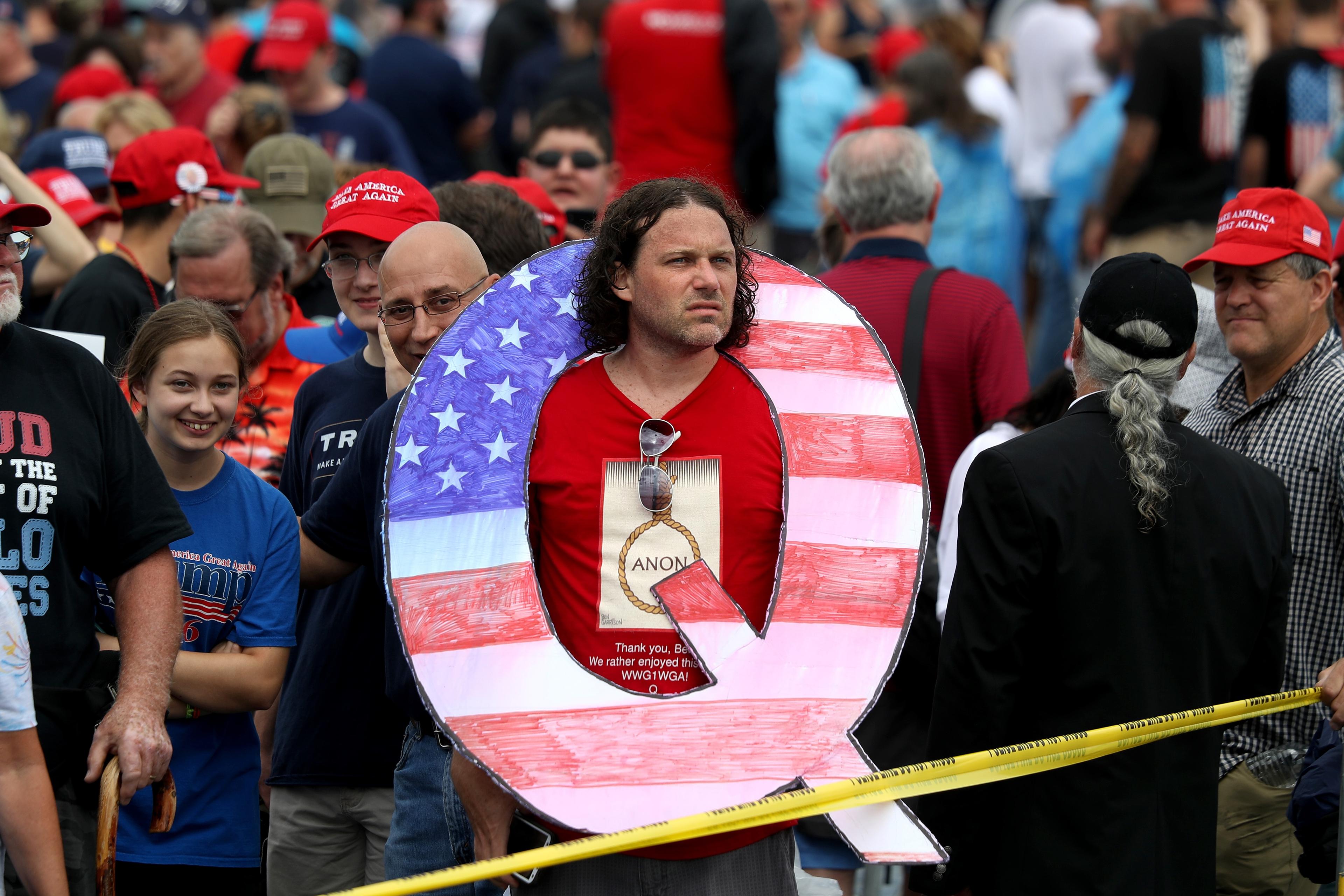 Photo of a man in a crowd wearing a large letter Q with the American flag pattern. Many people wear red hats.