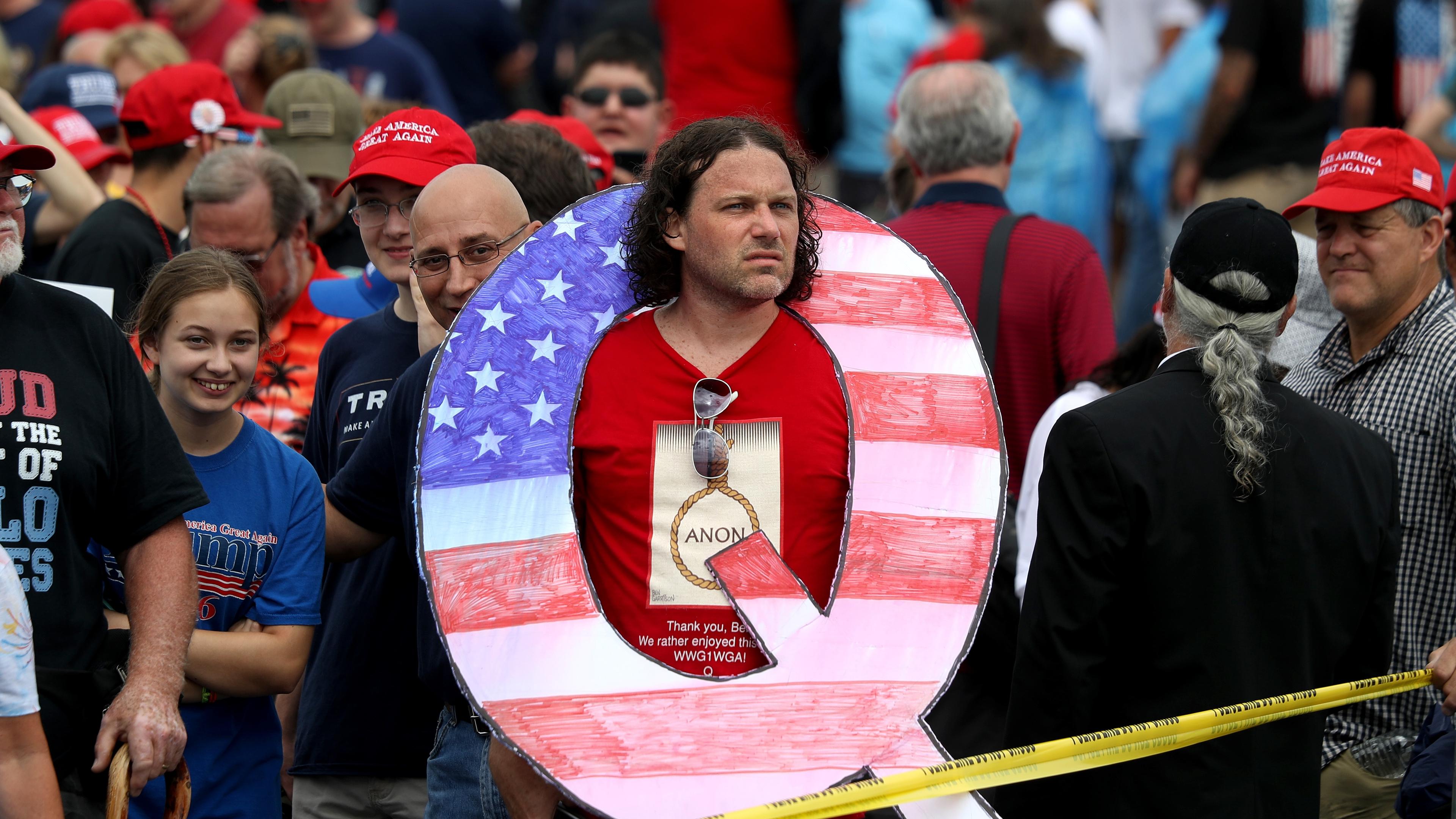Photo of a man in a crowd wearing a large letter Q with the American flag pattern. Many people wear red hats.