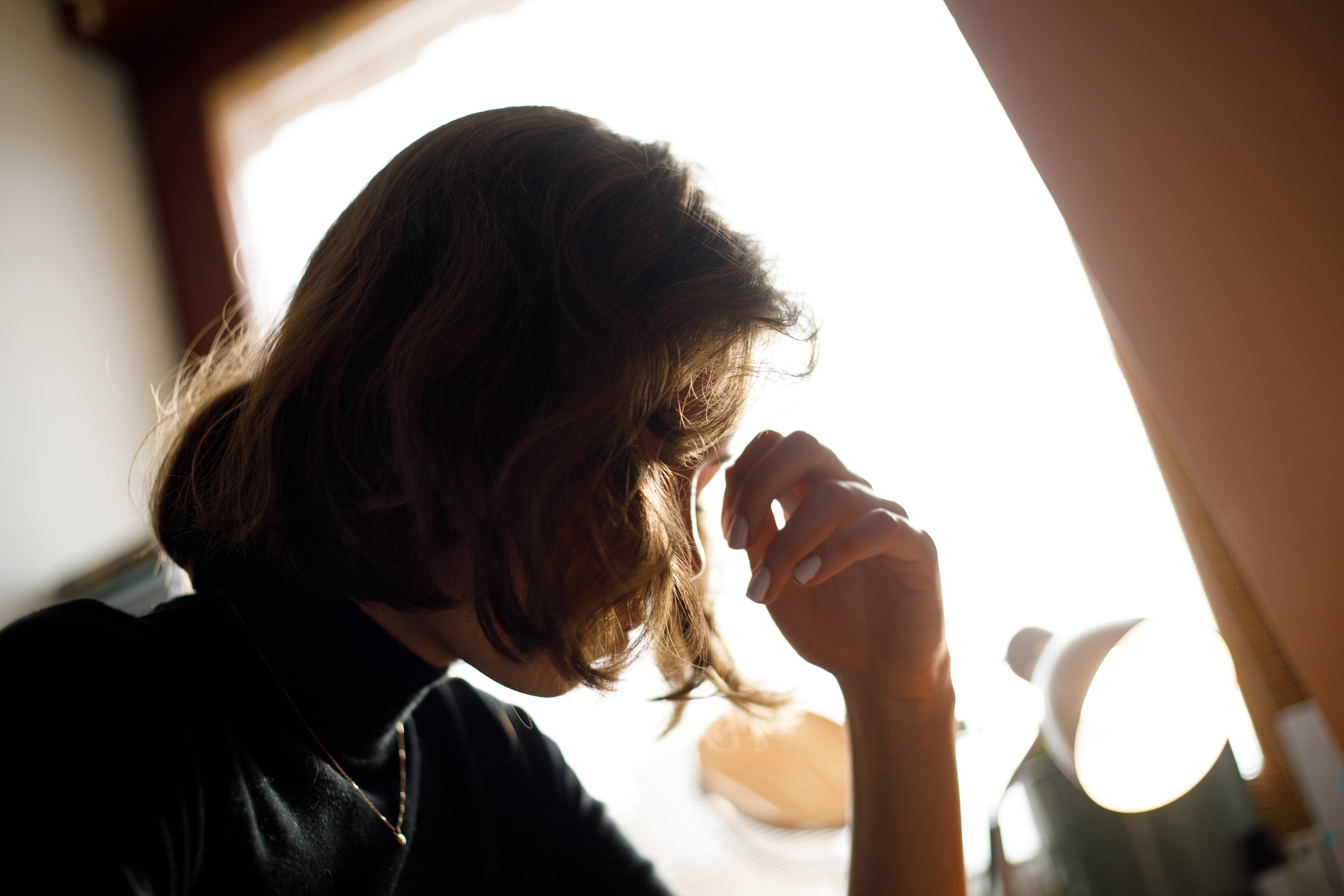 Photo of a person with shoulder-length hair in profile, sitting near a bright window, resting their head on their hand.