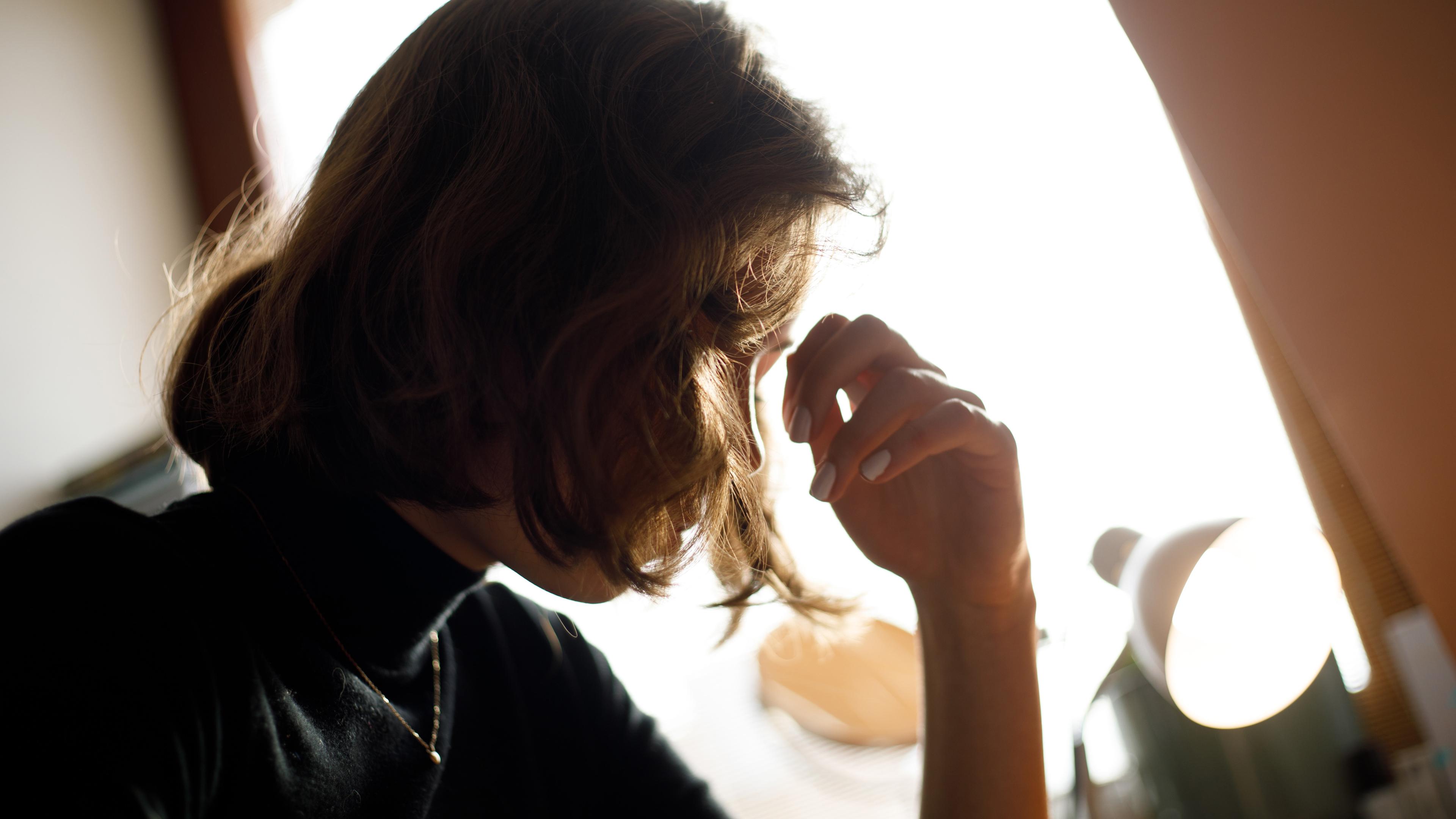 Photo of a person with shoulder-length hair in profile, sitting near a bright window, resting their head on their hand.