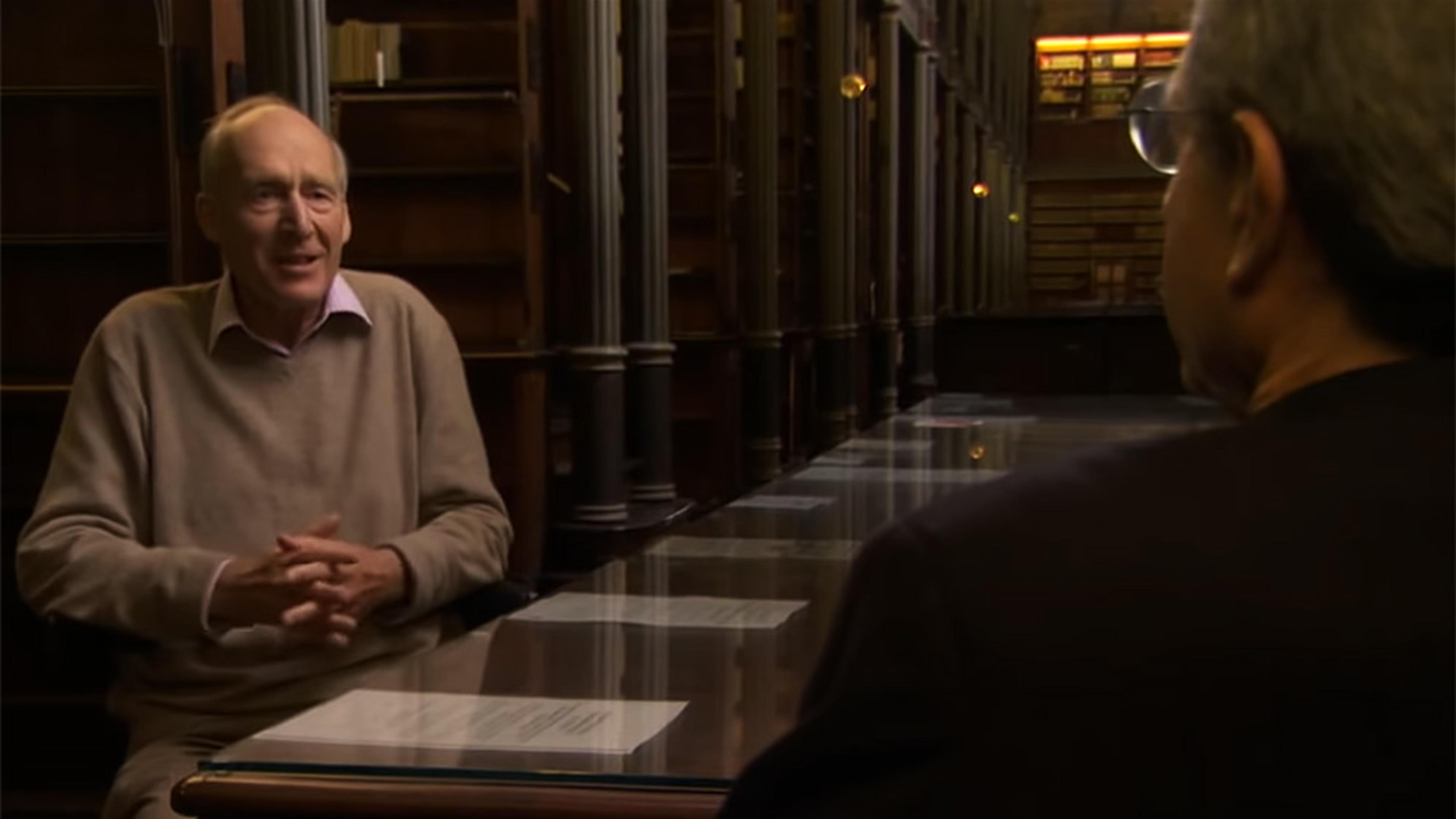 Two men conversing at a table in a library with tall shelves and warm lighting; one older man faces the camera, his hands clasped.