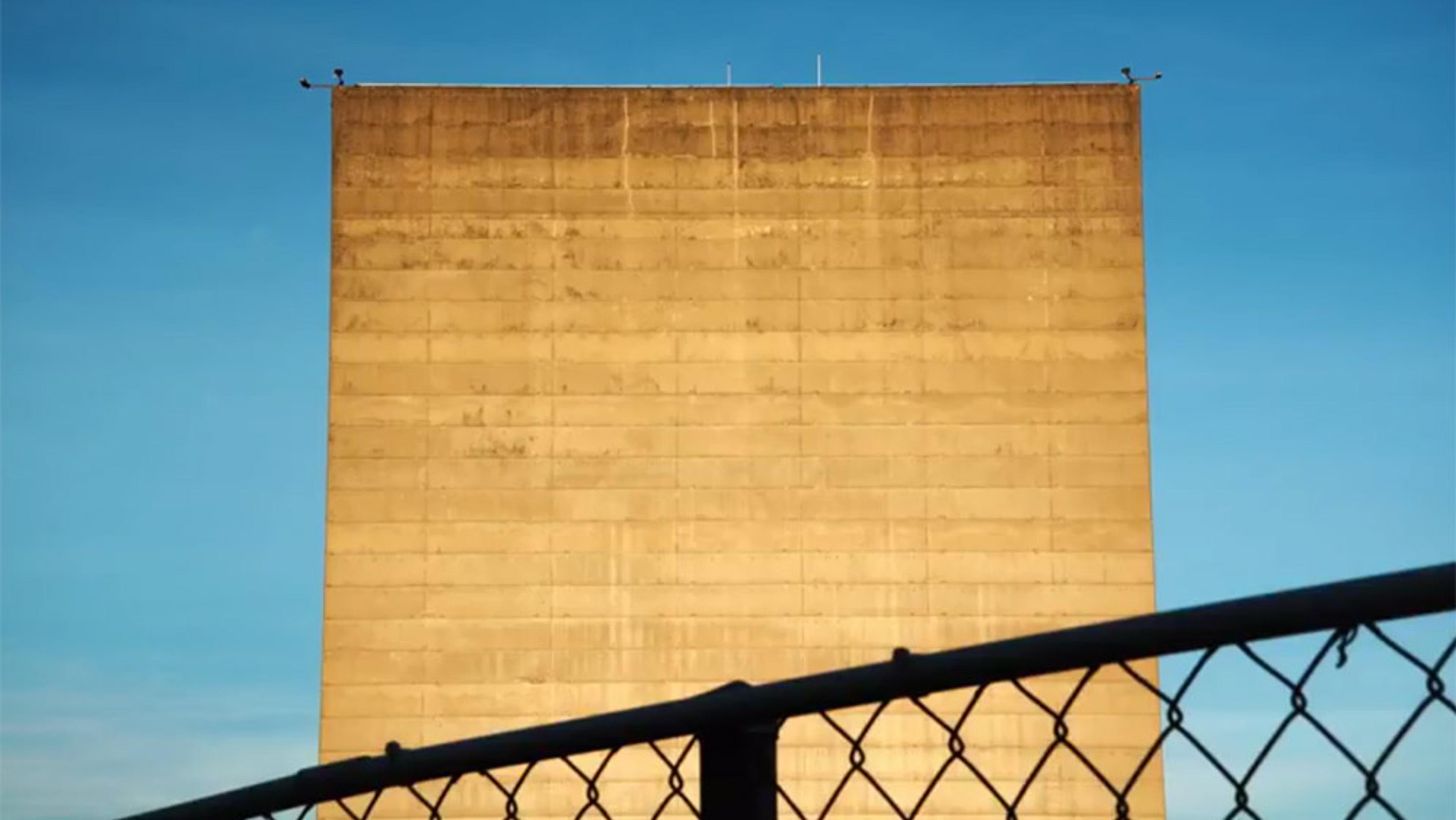 A tall, sunlit concrete building with a chain-link fence in the foreground against a clear blue sky.