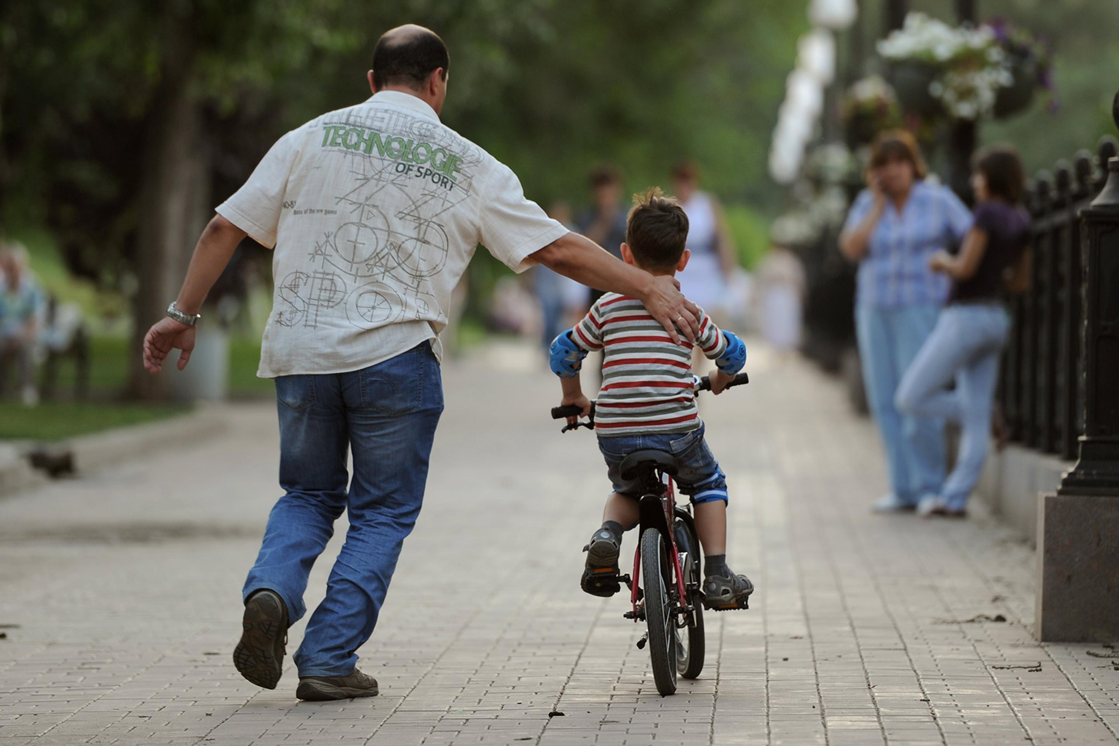 A man helps a child ride a bike on a paved walkway in a park. Other people are visible in the background.