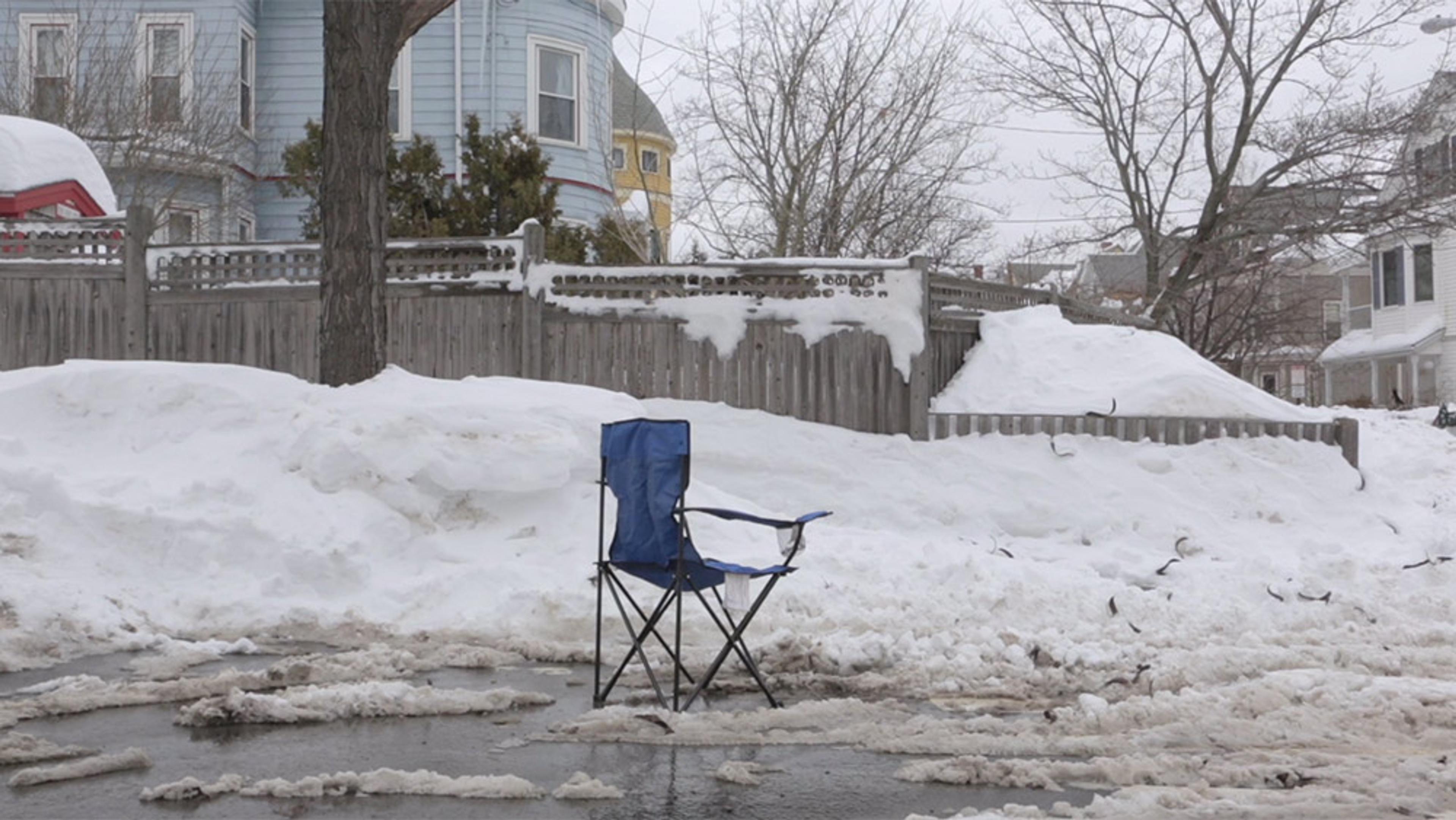 A snowy urban scene with a blue camping chair placed in the street, in front of a snow-covered fence and houses.