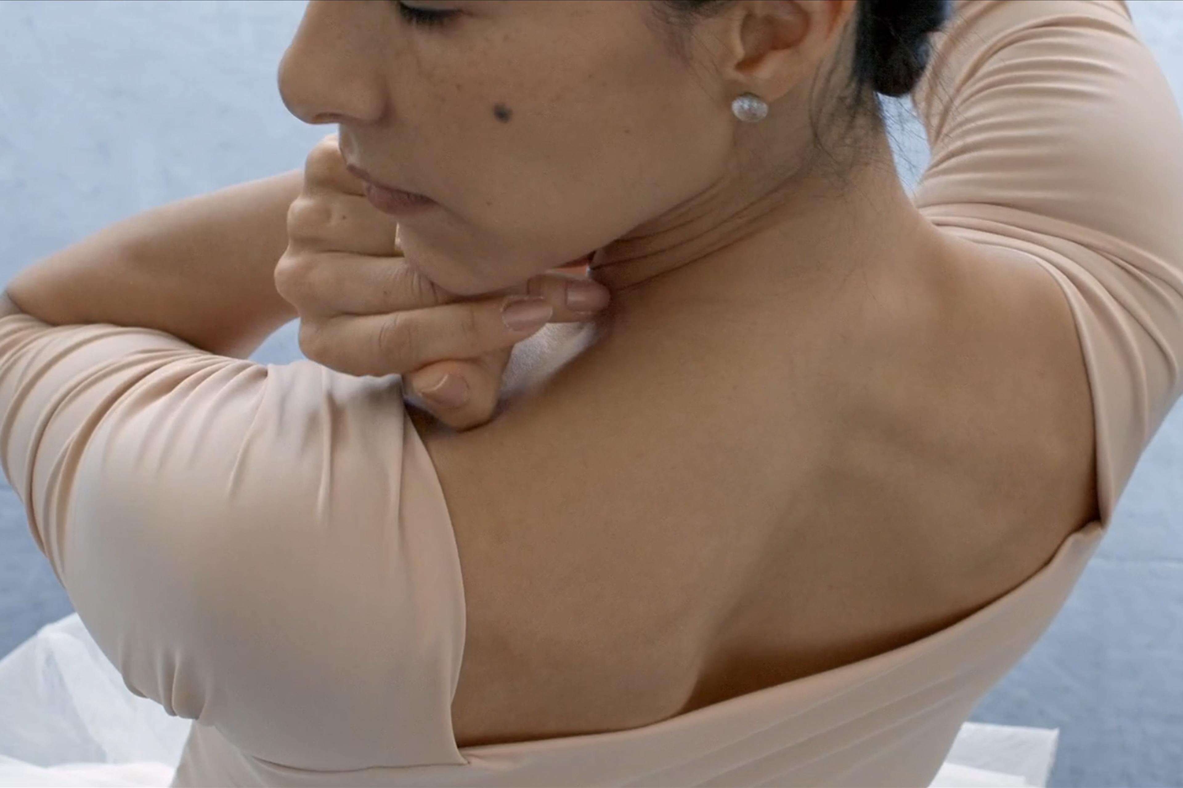 Close-up photo of a woman in a beige top with hand on neck showing her back and shoulder against a light blue background.
