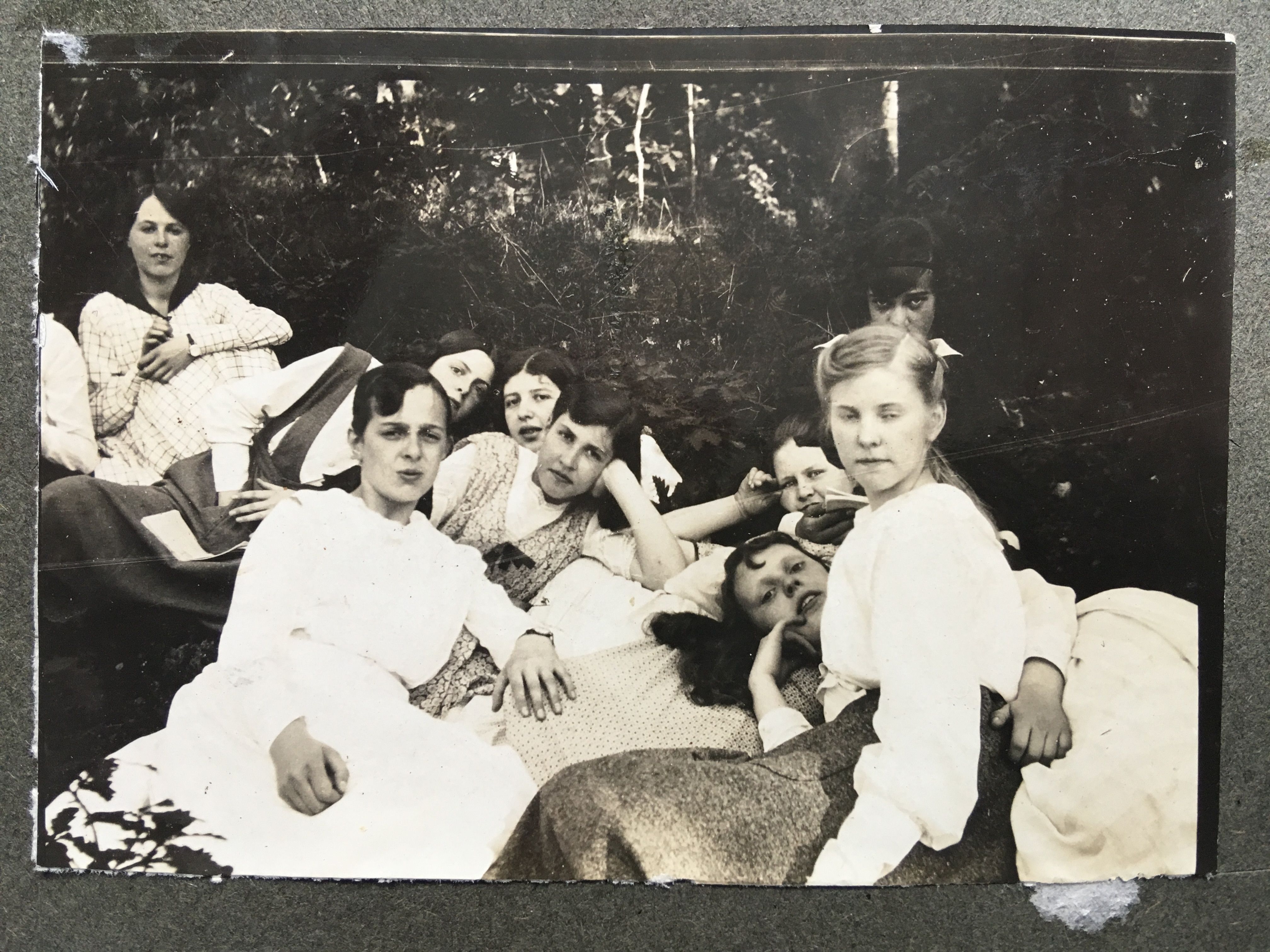Vintage photo of a group of women in white dresses lounging outdoors on the grass, with trees in the background.