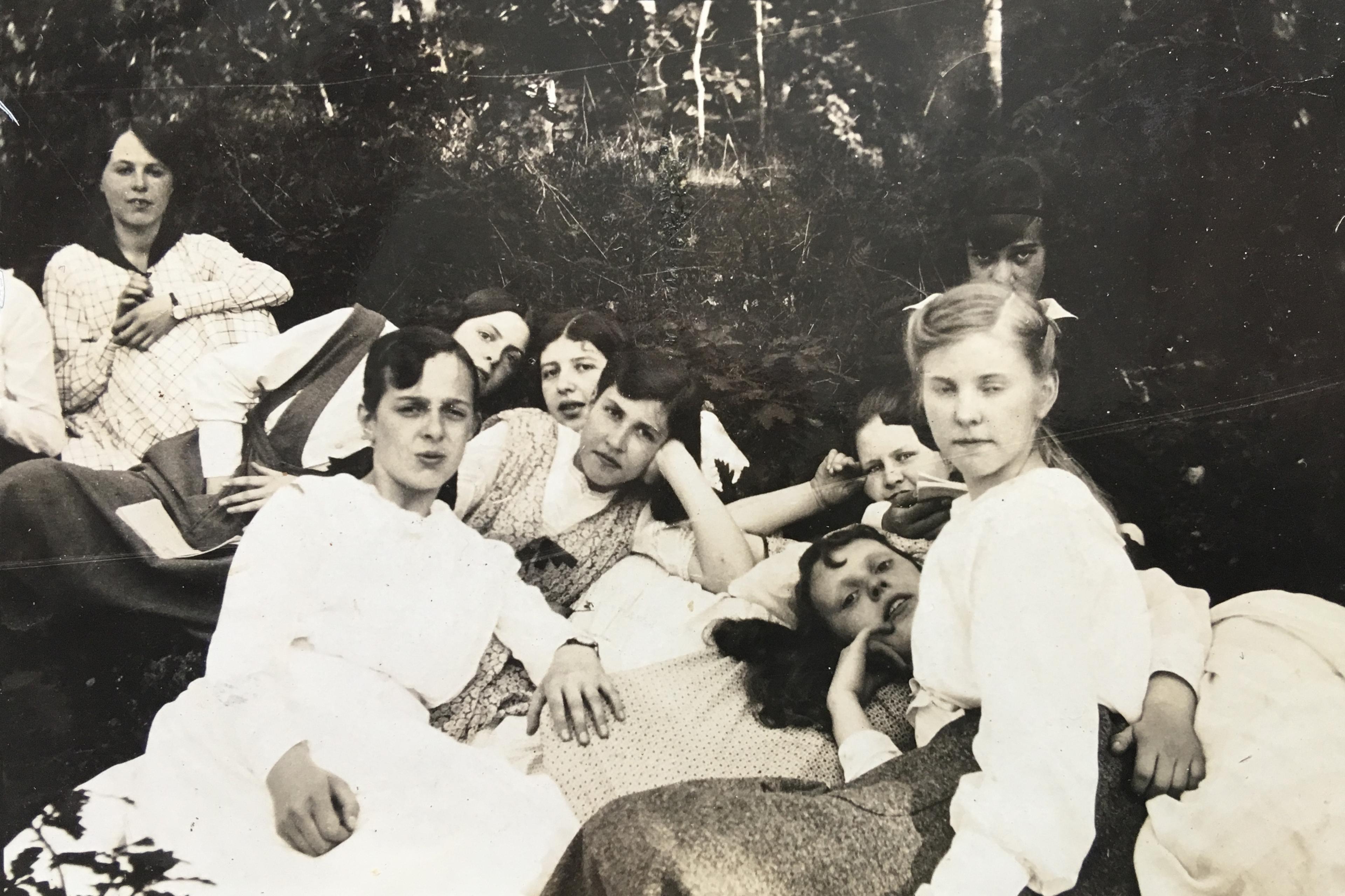Vintage photo of a group of women in white dresses lounging outdoors on the grass, with trees in the background.