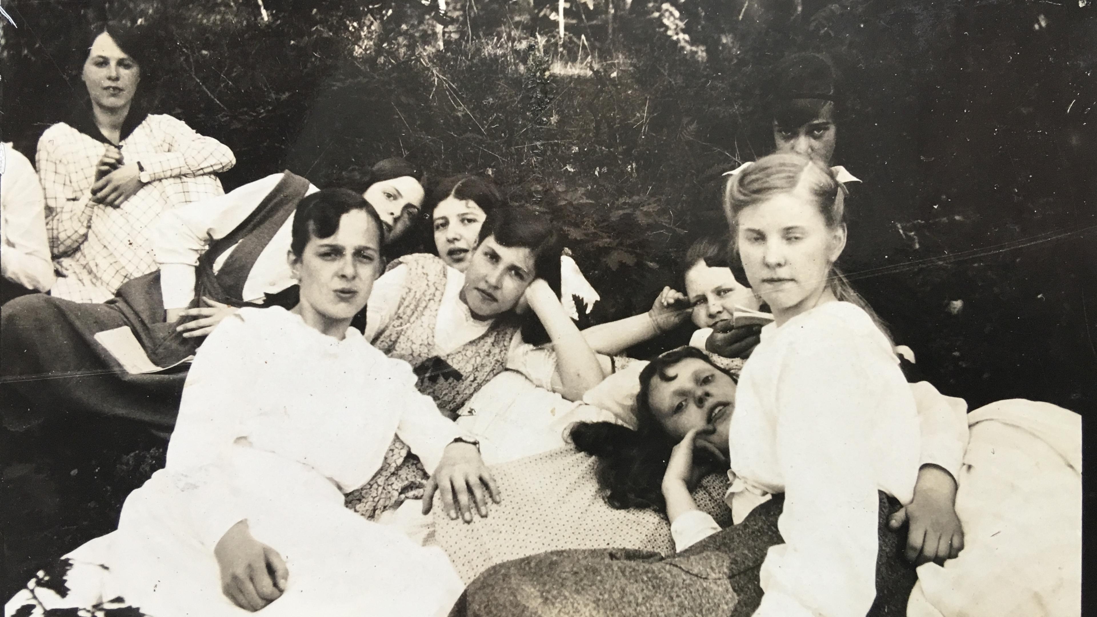 Vintage photo of a group of women in white dresses lounging outdoors on the grass, with trees in the background.