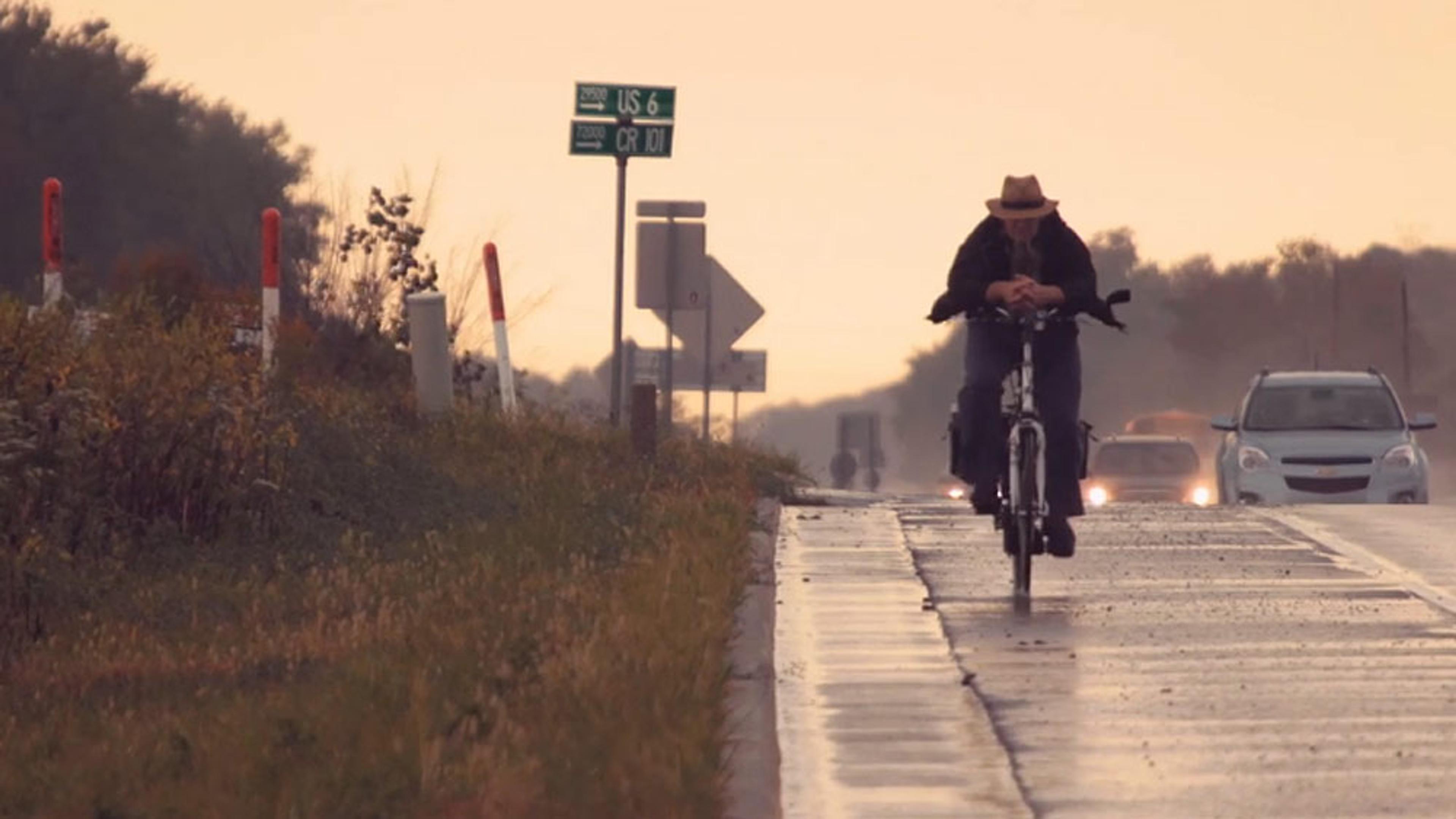 A person biking on a wet road near a grassy verge with cars and signposts visible in the background under an overcast sky.