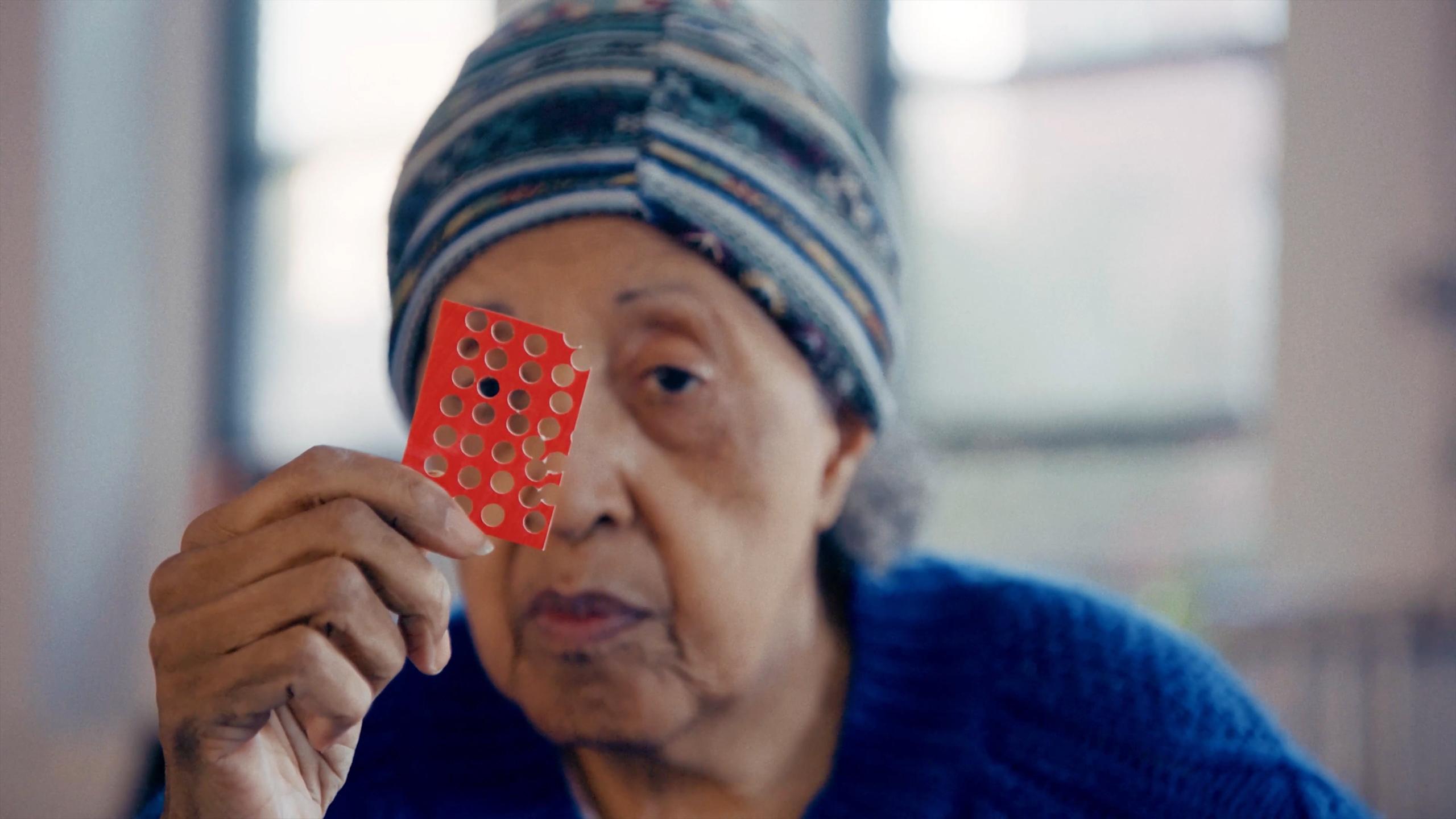 An elderly woman in a patterned beanie holding a red card with holes punched into it in front of her face.