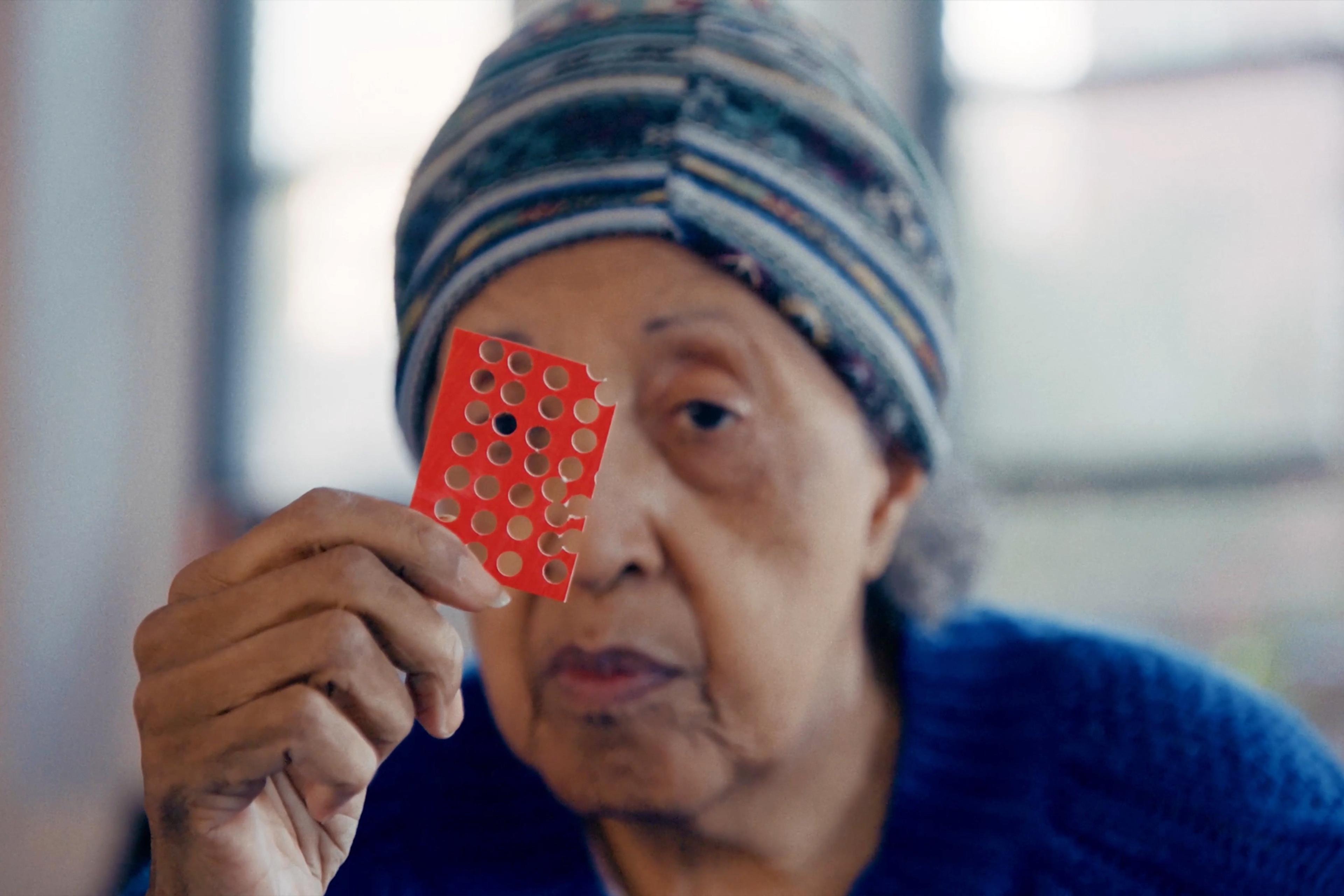 An elderly woman in a patterned beanie holding a red card with holes punched into it in front of her face.