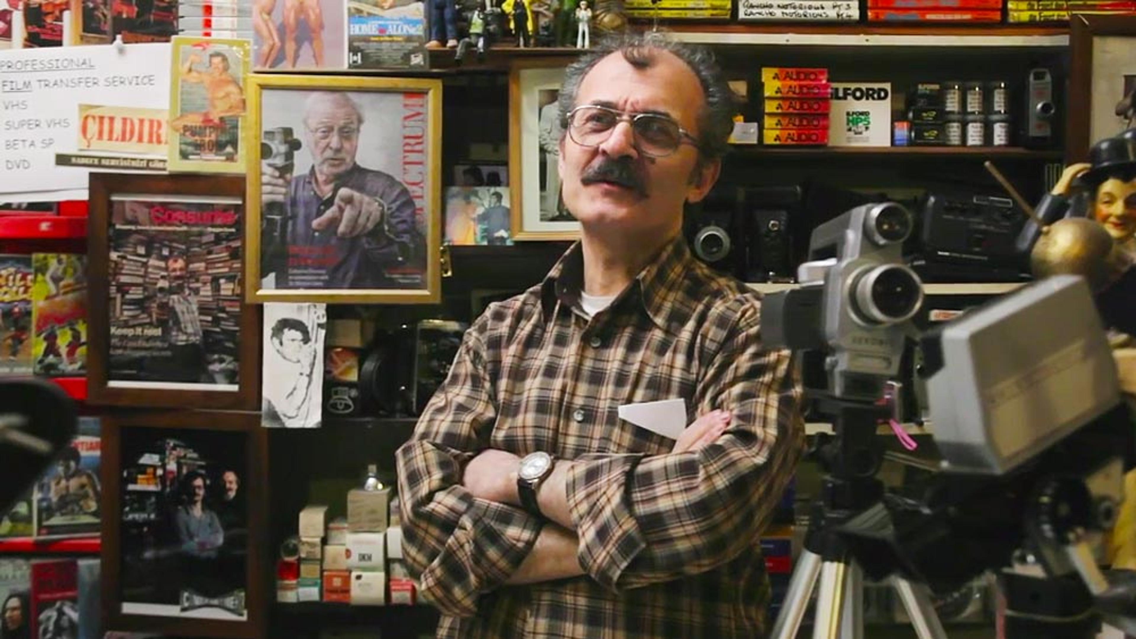 A man in a plaid shirt with glasses and a moustache standing in a cluttered room filled with cameras and film equipment.