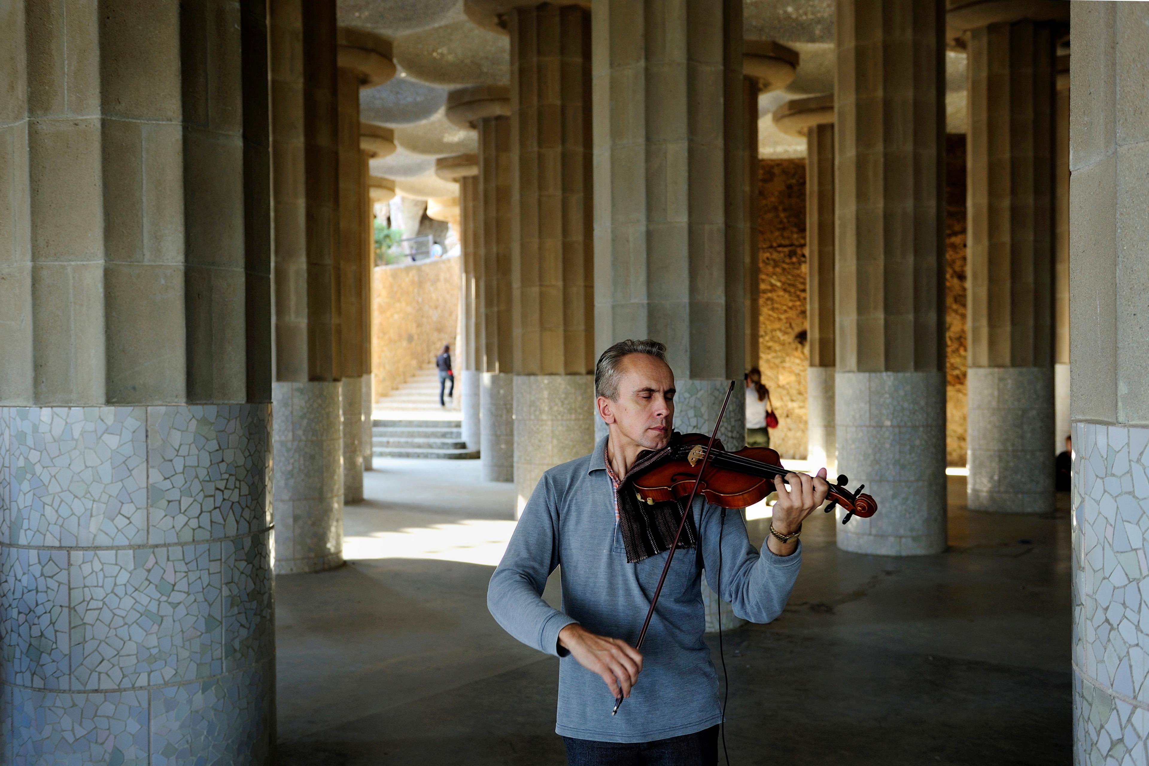 Photo of a man playing a violin in a columned, mosaic-tiled hall with people walking in the background.
