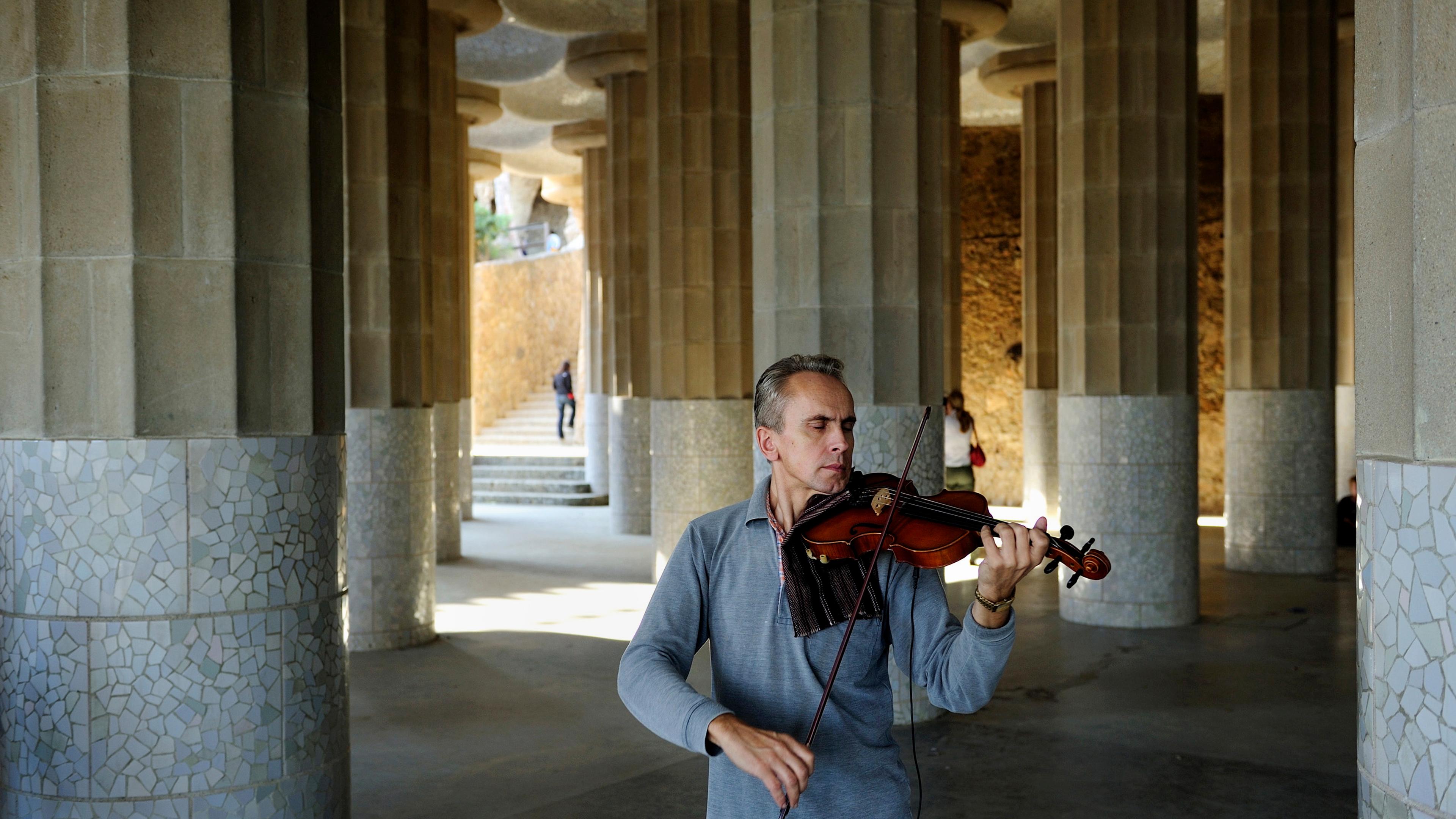Photo of a man playing a violin in a columned, mosaic-tiled hall with people walking in the background.