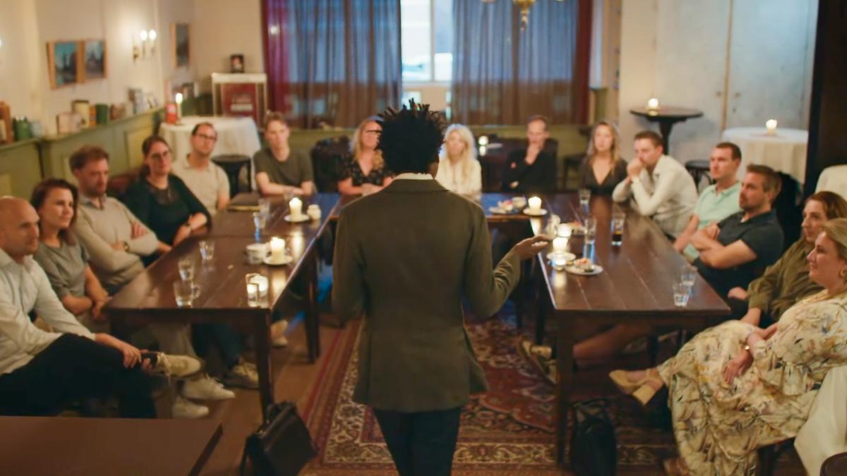 Photo of a man speaking to a group seated around tables in a candlelit room, view from behind the speaker.