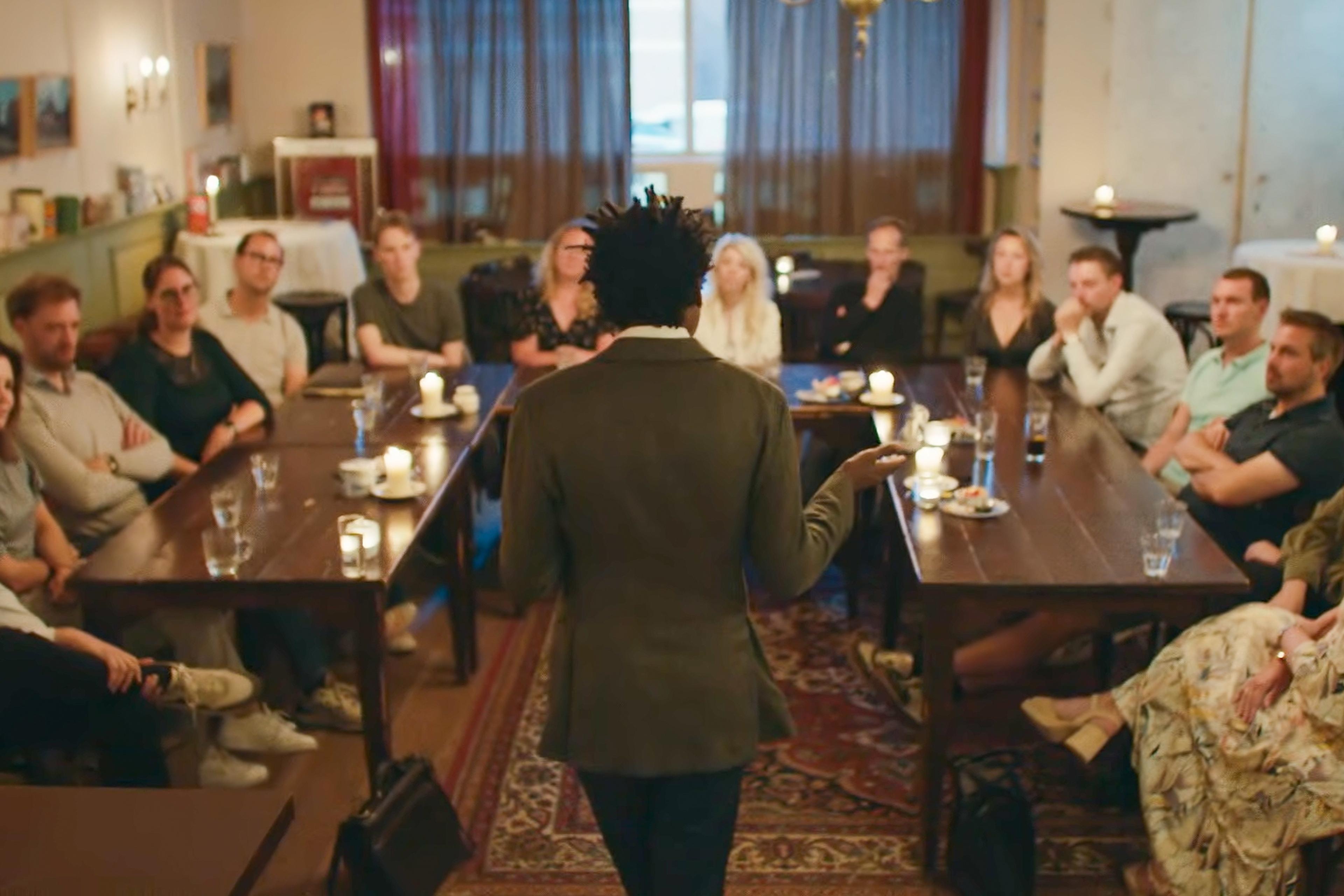 Photo of a man speaking to a group seated around tables in a candlelit room, view from behind the speaker.