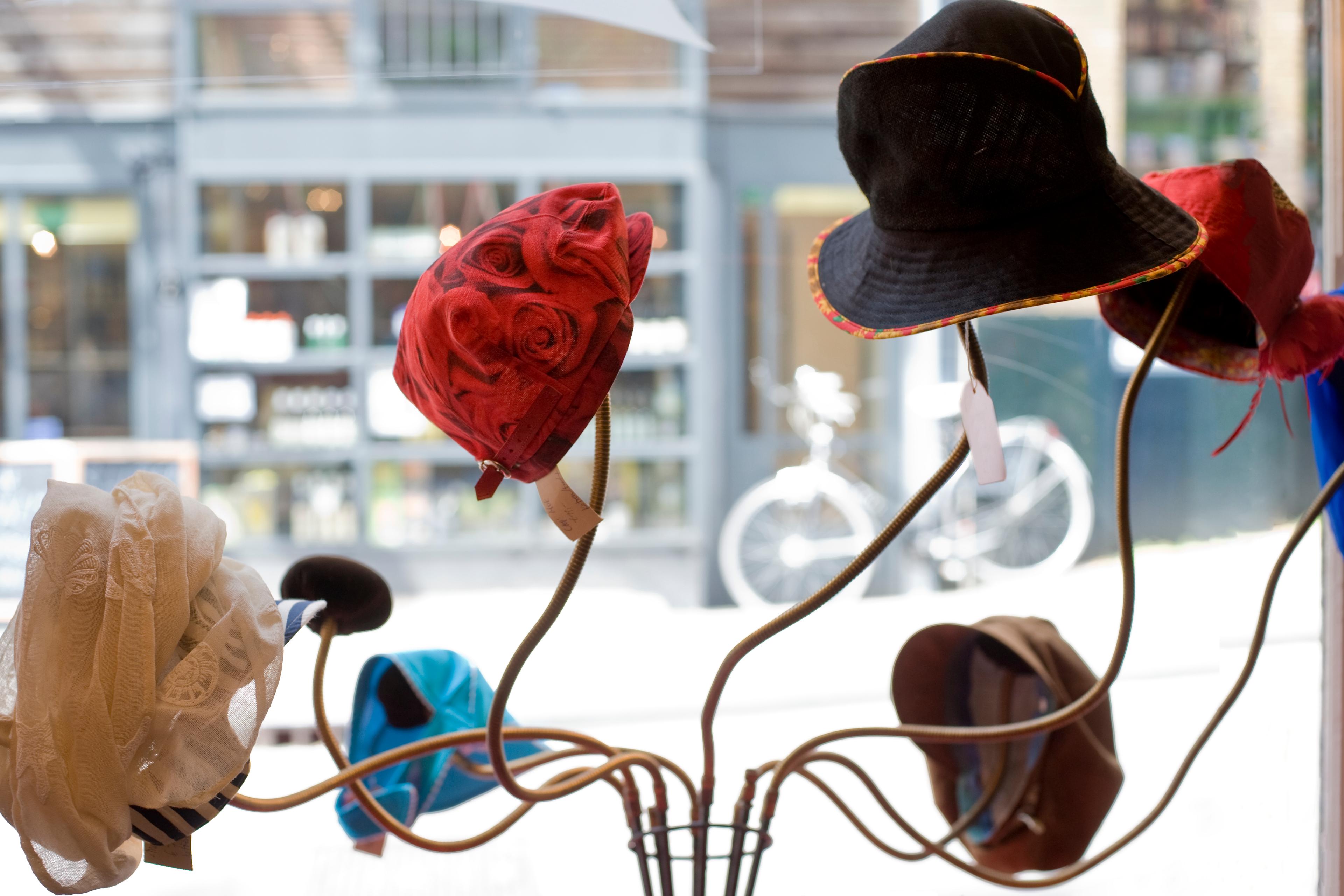 Various hats displayed on stands in a shop window with a blurred street scene and bicycle outside.