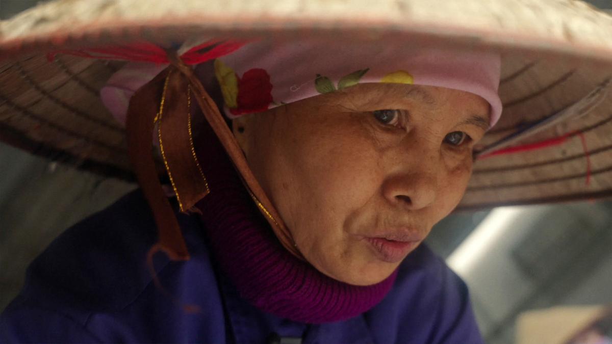 Photo of a woman wearing a traditional conical hat and purple clothing, with a floral scarf visible beneath the hat.