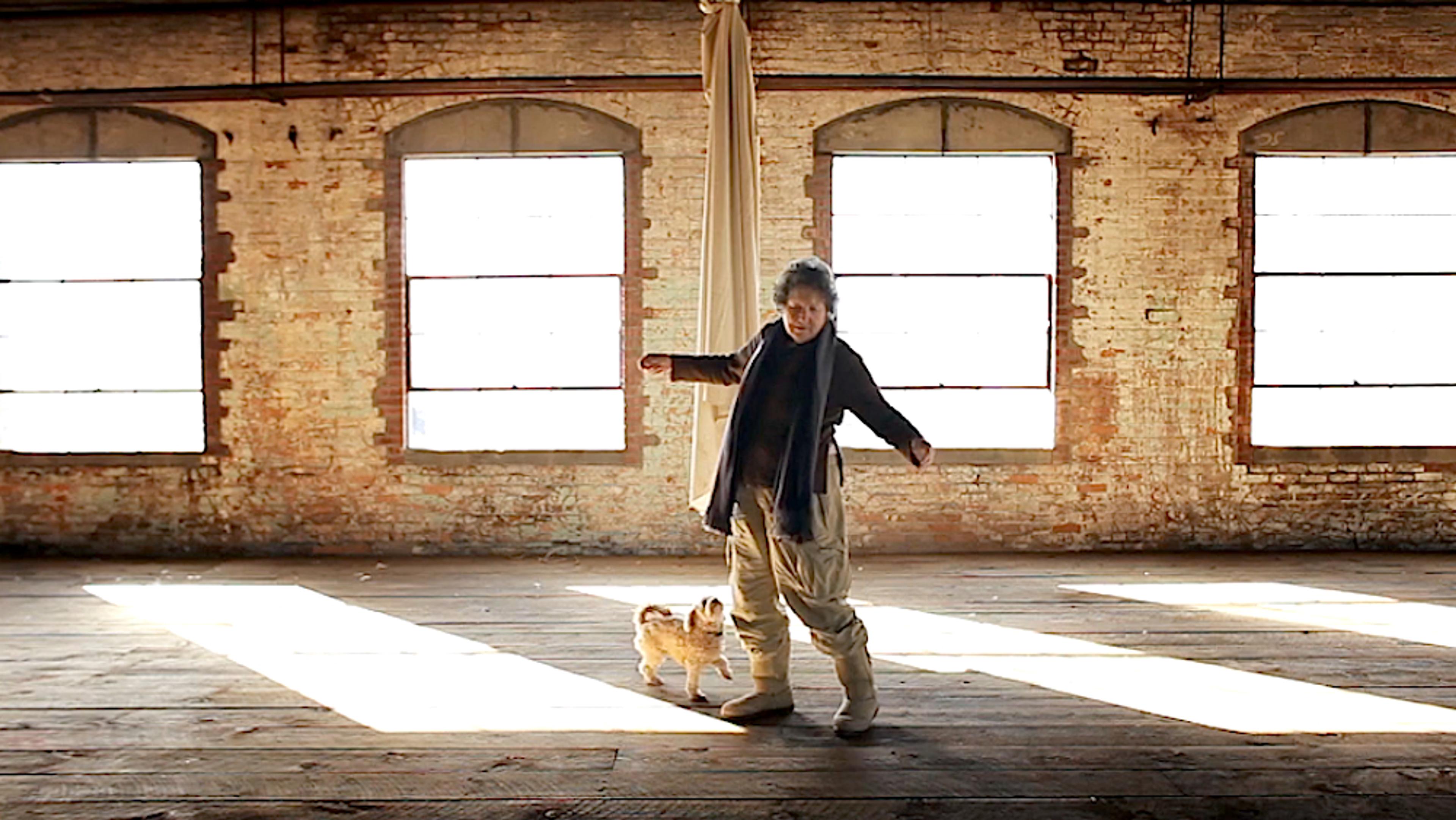 An elderly woman and a small dog standing in a large empty room with wooden floors and brick walls, illuminated by sunlight through windows.