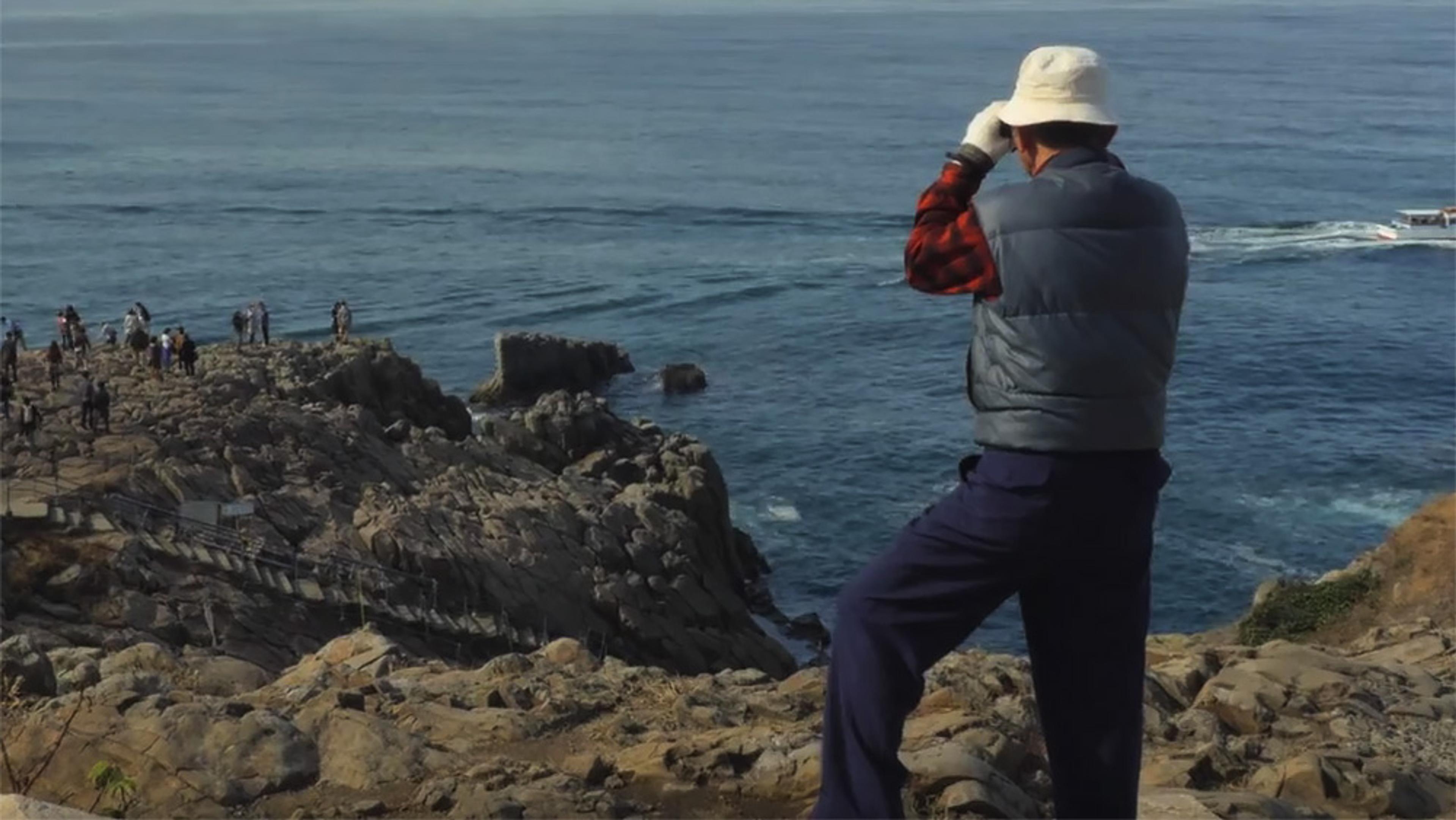 A person in a hat looking at the sea atop rocky cliffs, with other people in the distance and a boat on the water.