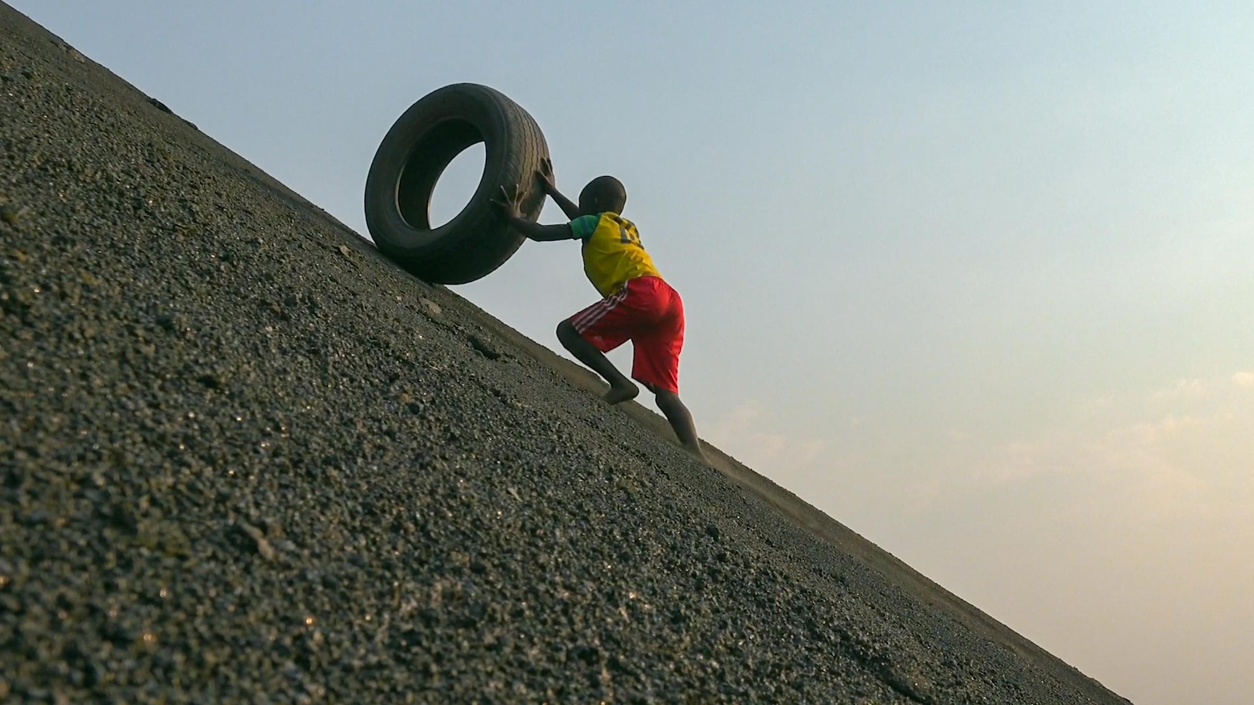 A child in a yellow shirt and red shorts pushing a tire uphill on a steep, gravelly incline against a clear sky backdrop.