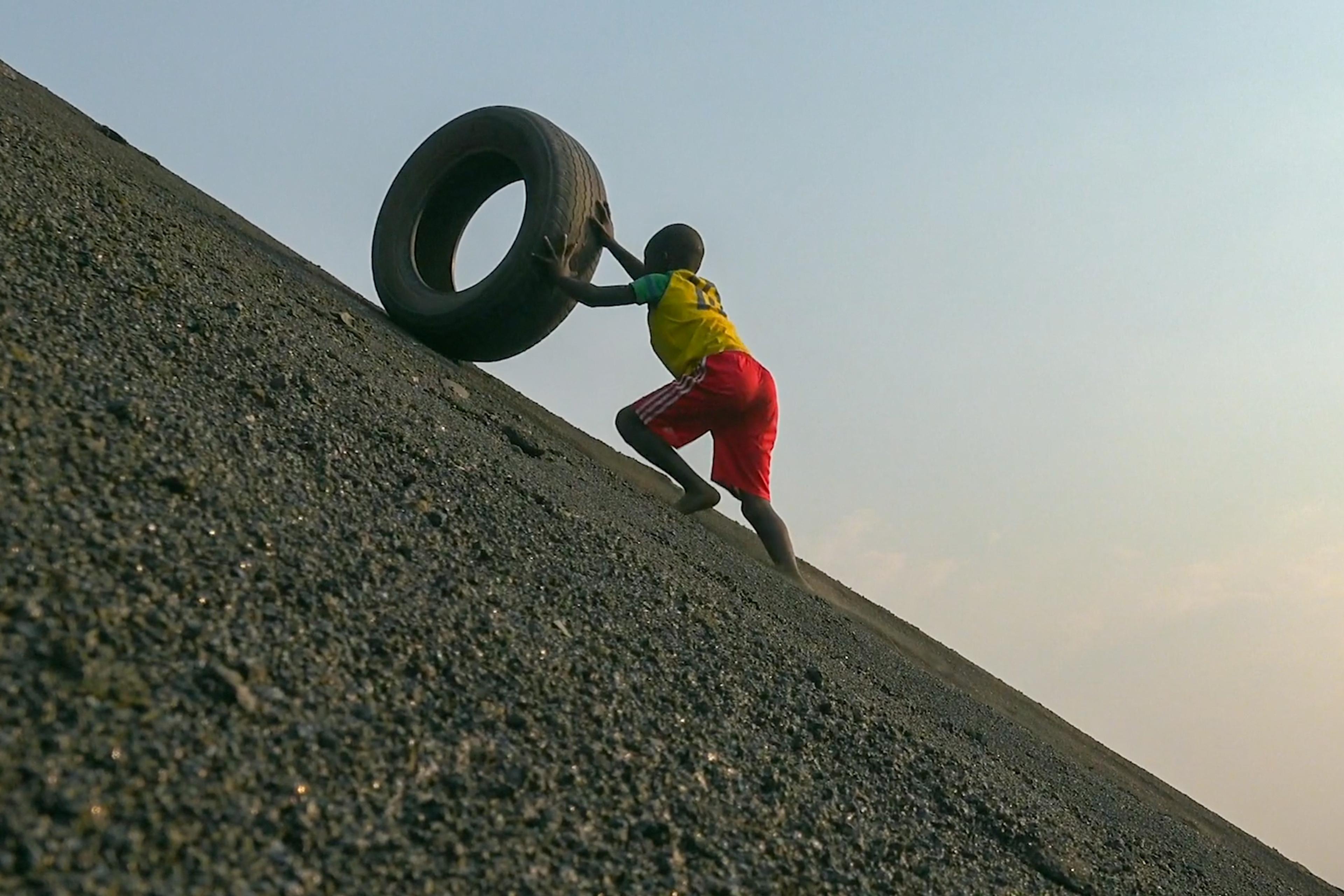 A child in a yellow shirt and red shorts pushing a tire uphill on a steep, gravelly incline against a clear sky backdrop.