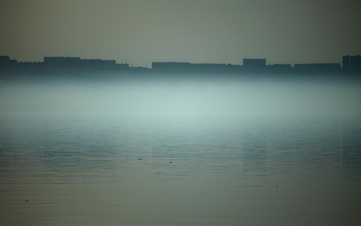 Mist-covered city skyline with a calm, reflective body of water in the foreground under a grey sky.