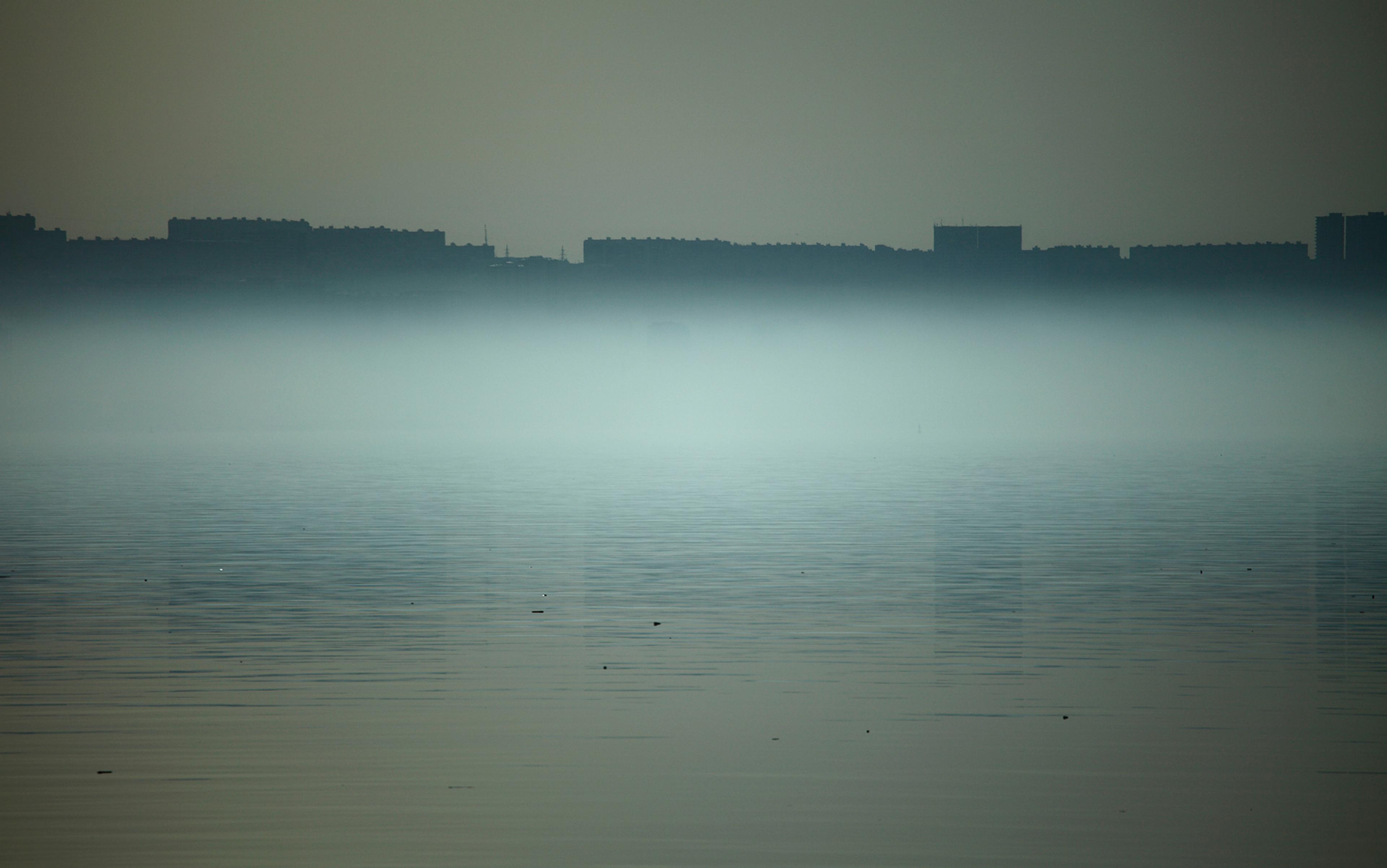 Mist-covered city skyline with a calm, reflective body of water in the foreground under a grey sky.