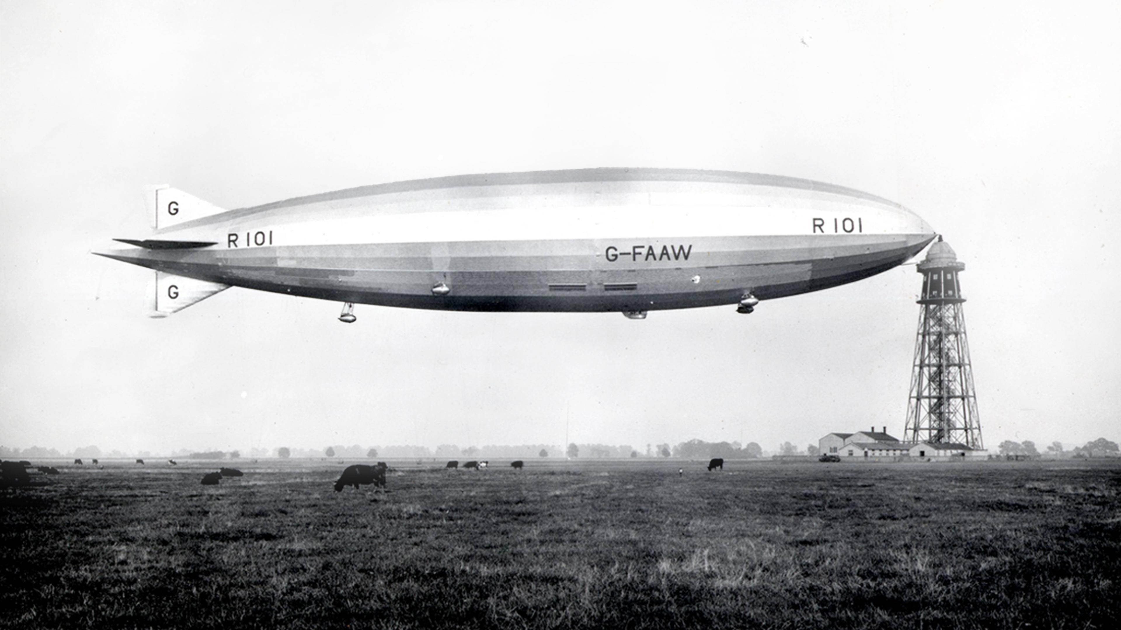 Photo of an airship labelled R101 flying above a grassy field with cows and a tall structure in the background.