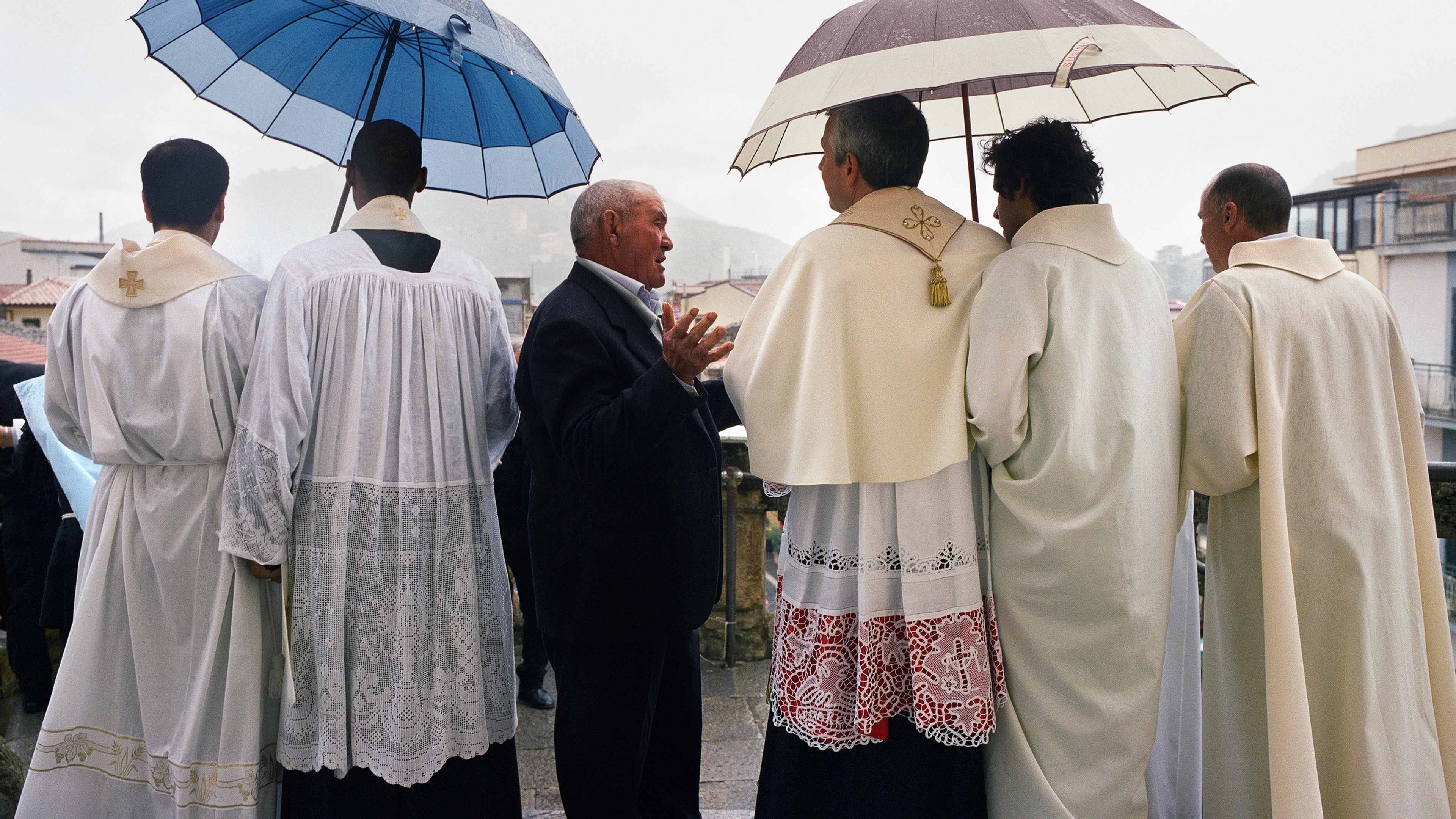 Photo of clergy in white robes and a man with umbrellas, standing outdoors on a rainy day.